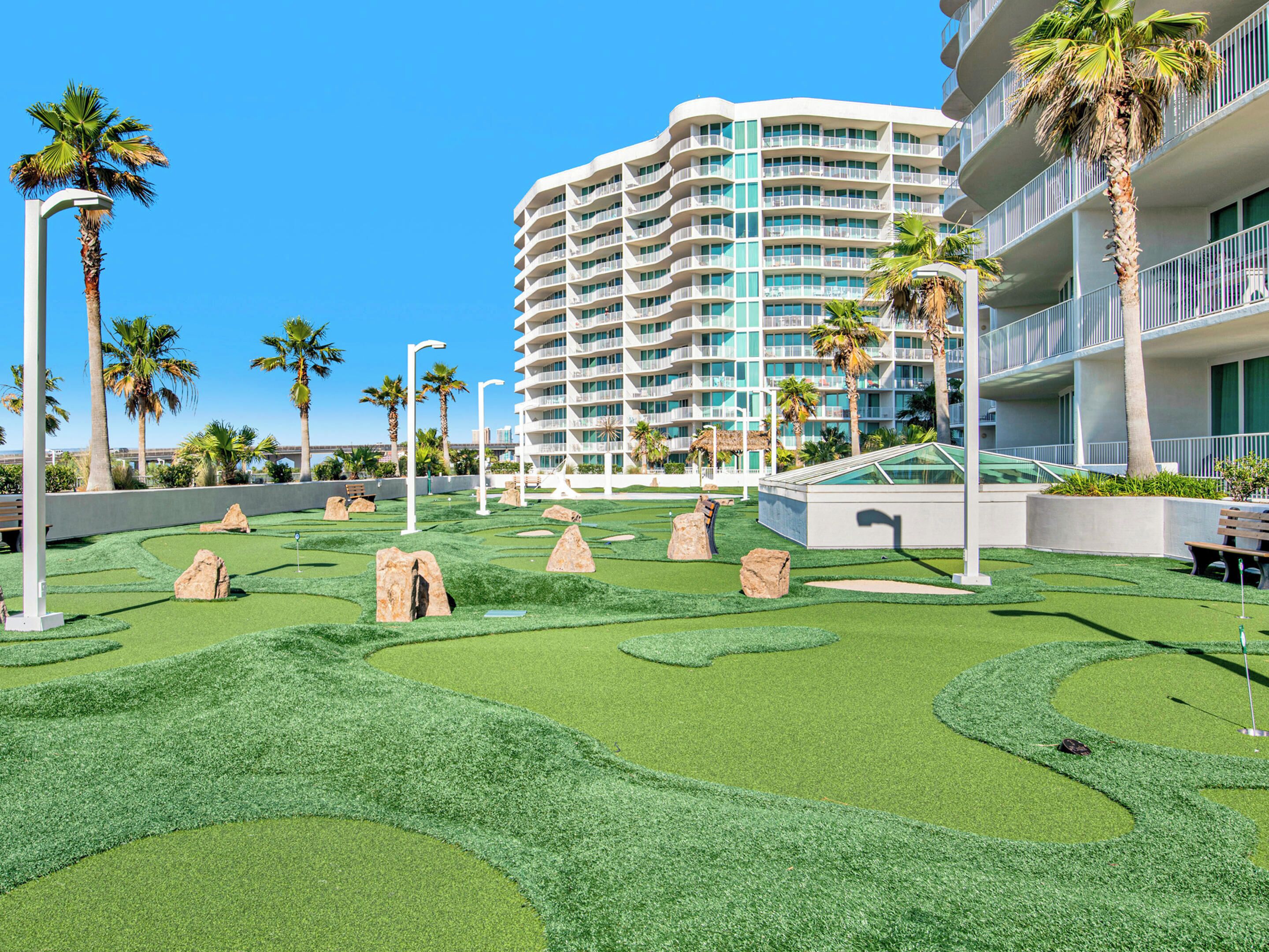 A modern hotel with a curved facade, multiple balconies, and a well-maintained artificial lawn with rock landscaping.