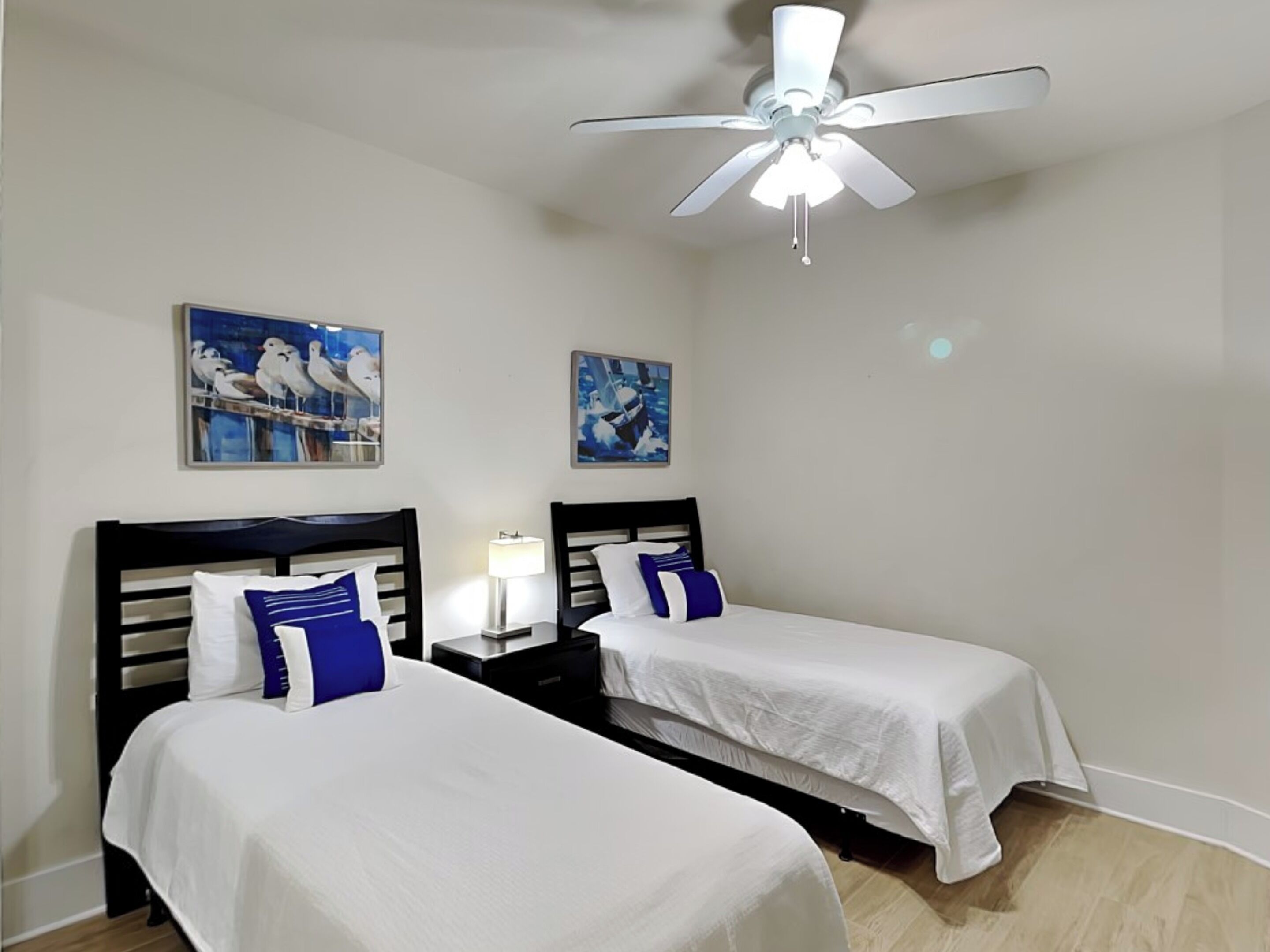 A hotel room with two single beds, a ceiling fan, and two wall-mounted paintings.