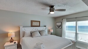 A neatly made bed with white bedding and two pillows, a bedside table with a lamp, a ceiling fan, and a window with a view of the ocean. - Sea Oats #603 by Southern Vacation Rentals (Fort Walton Beach)