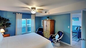Un dormitorio con cama, ventilador de techo, armario de madera, una silla y vistas a una habitación azul a través de una puerta abierta.