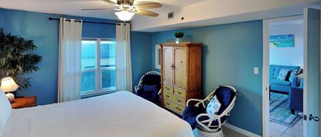 A bedroom with a bed, a ceiling fan, a wooden wardrobe, a chair, and a view of a blue room through an open door.