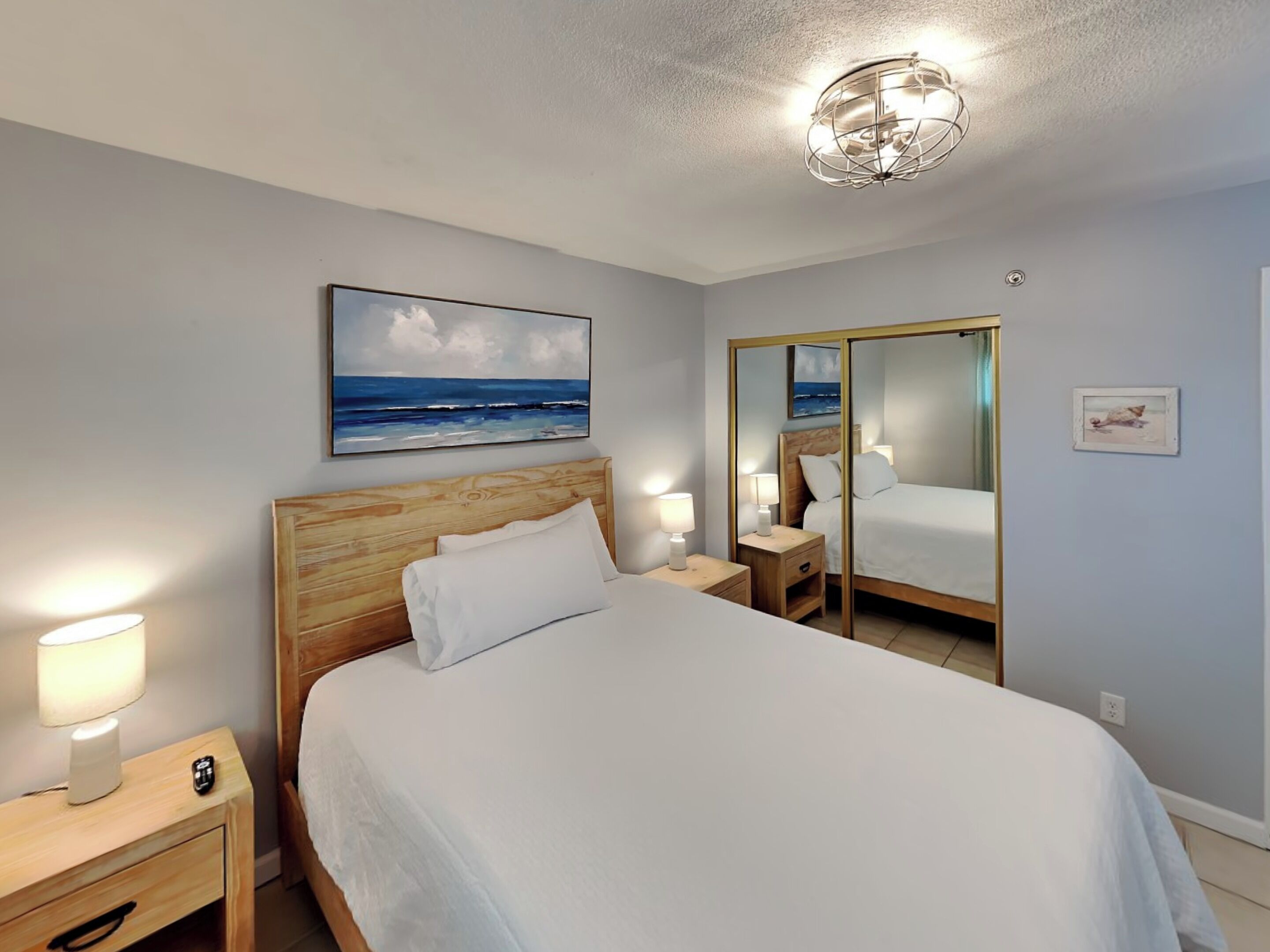 A bedroom with a wooden headboard, a large bed, bedside lamps, a nightstand, a mirror, and a painting of the ocean.