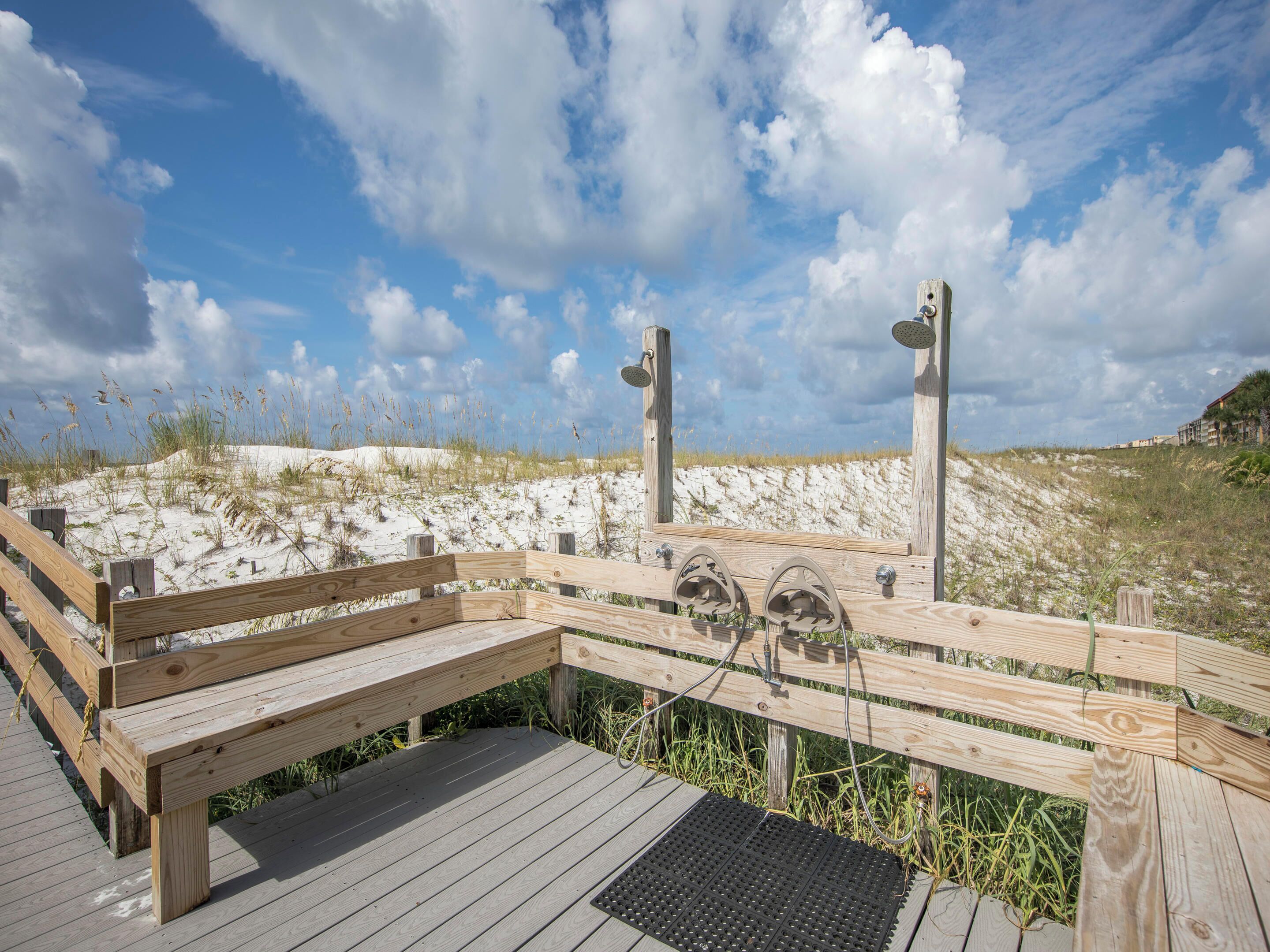 Een houten terras met een bankje en een zwarte afvoerrooster, met uitzicht op een zandstrand met duinen en een heldere lucht.