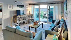 Sala de estar con un sofá, un sillón y un televisor sobre un mueble de madera. Hay una mesa con una lámpara y un jarrón. La habitación tiene vistas a un balcón con sillas y una mesa afuera.