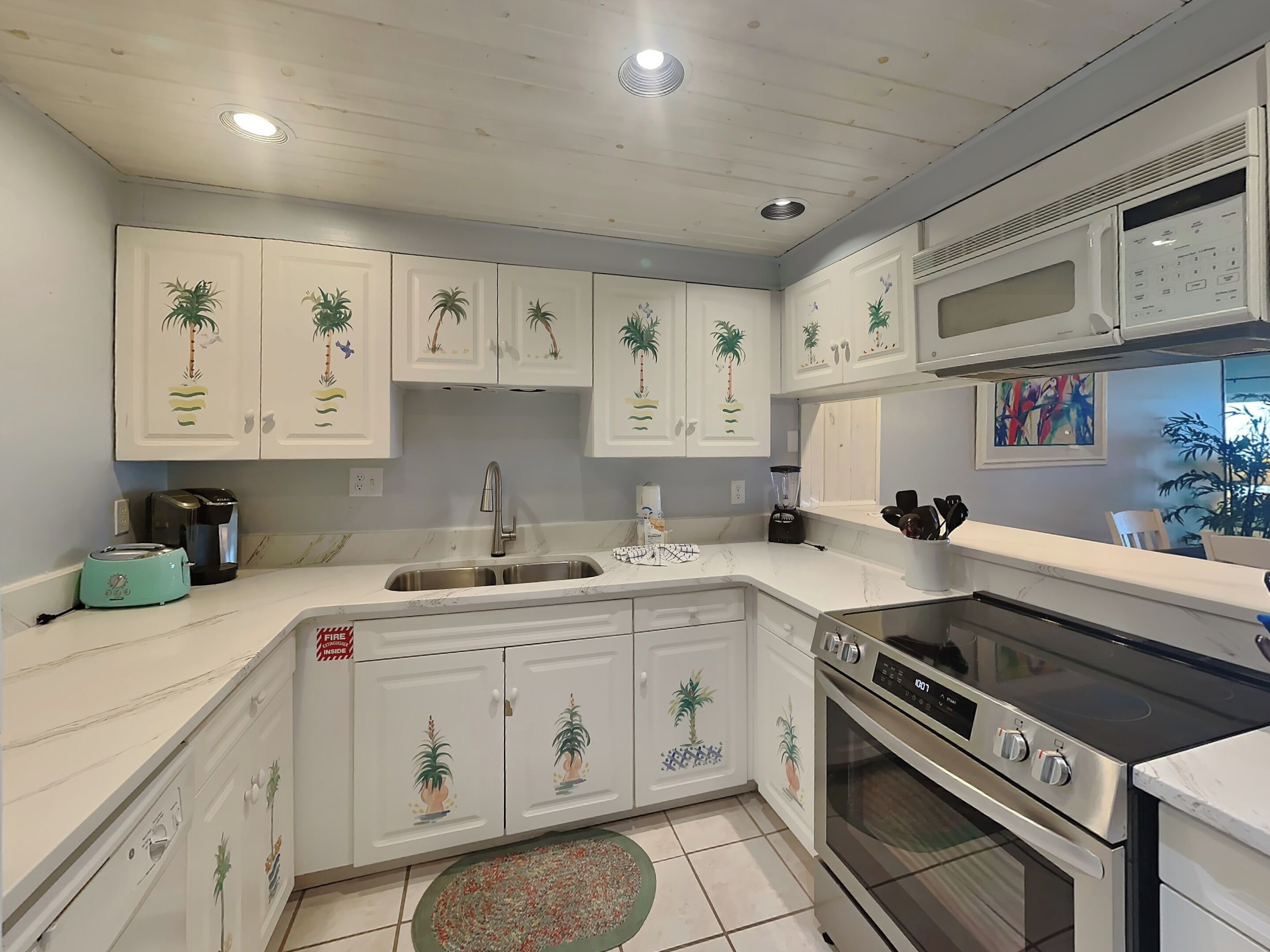 A kitchen with white cabinets featuring palm tree designs, a stainless steel sink, a toaster, a blender, and a stove.