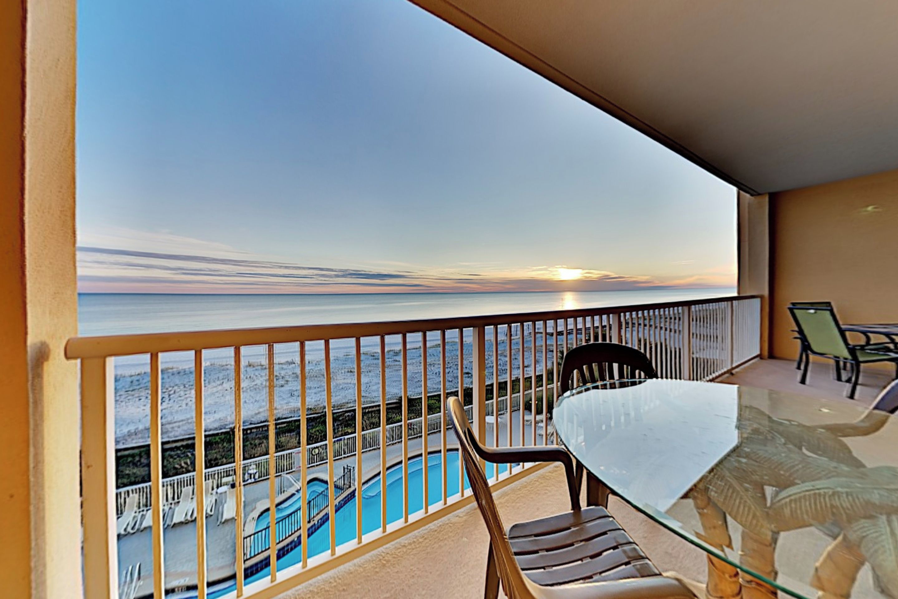 A balcony with a glass table and chairs, overlooking a swimming pool and a sunset view.