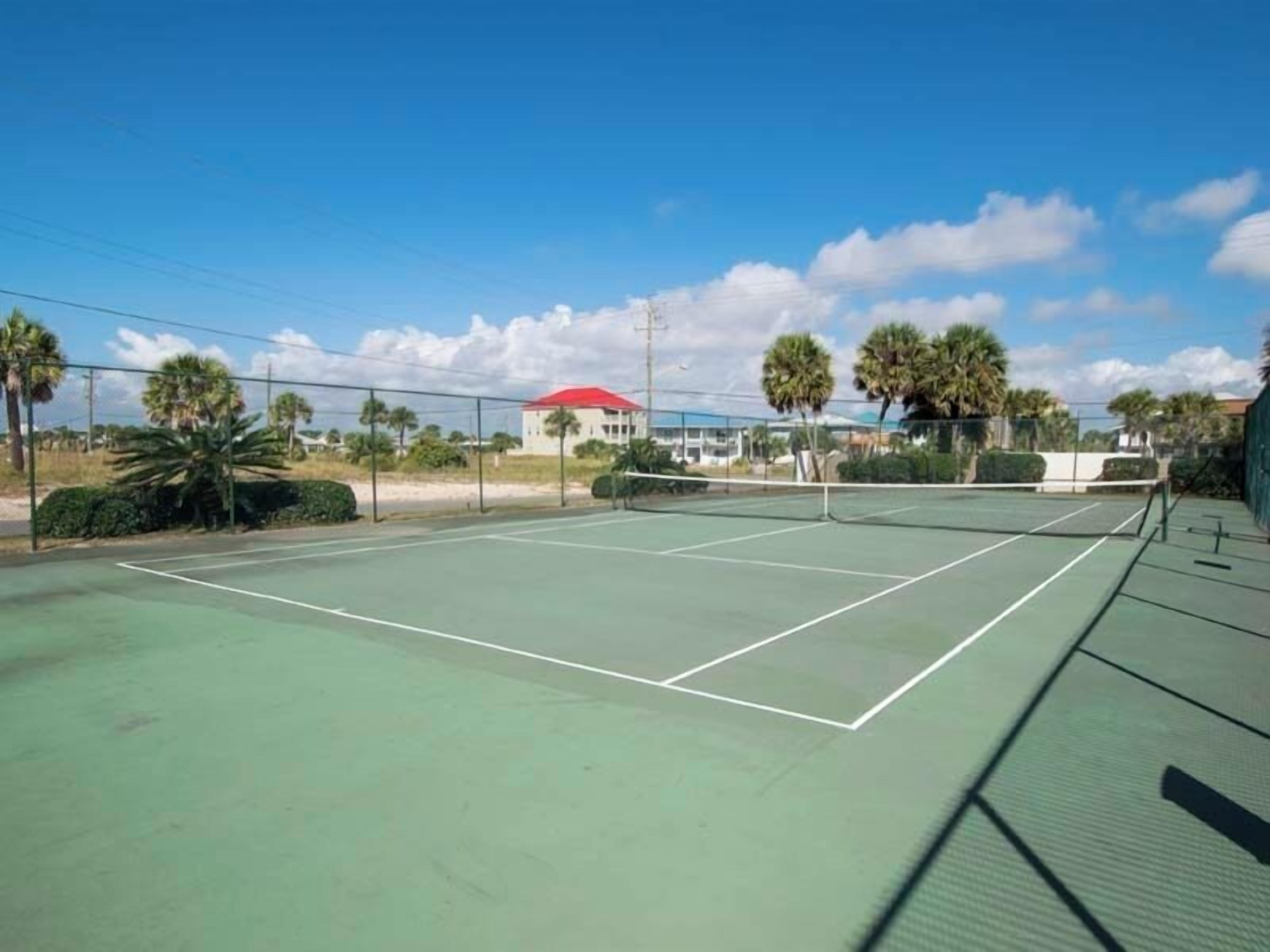 An outdoor tennis court with a green surface, surrounded by palm trees and a building with a red roof in the background.