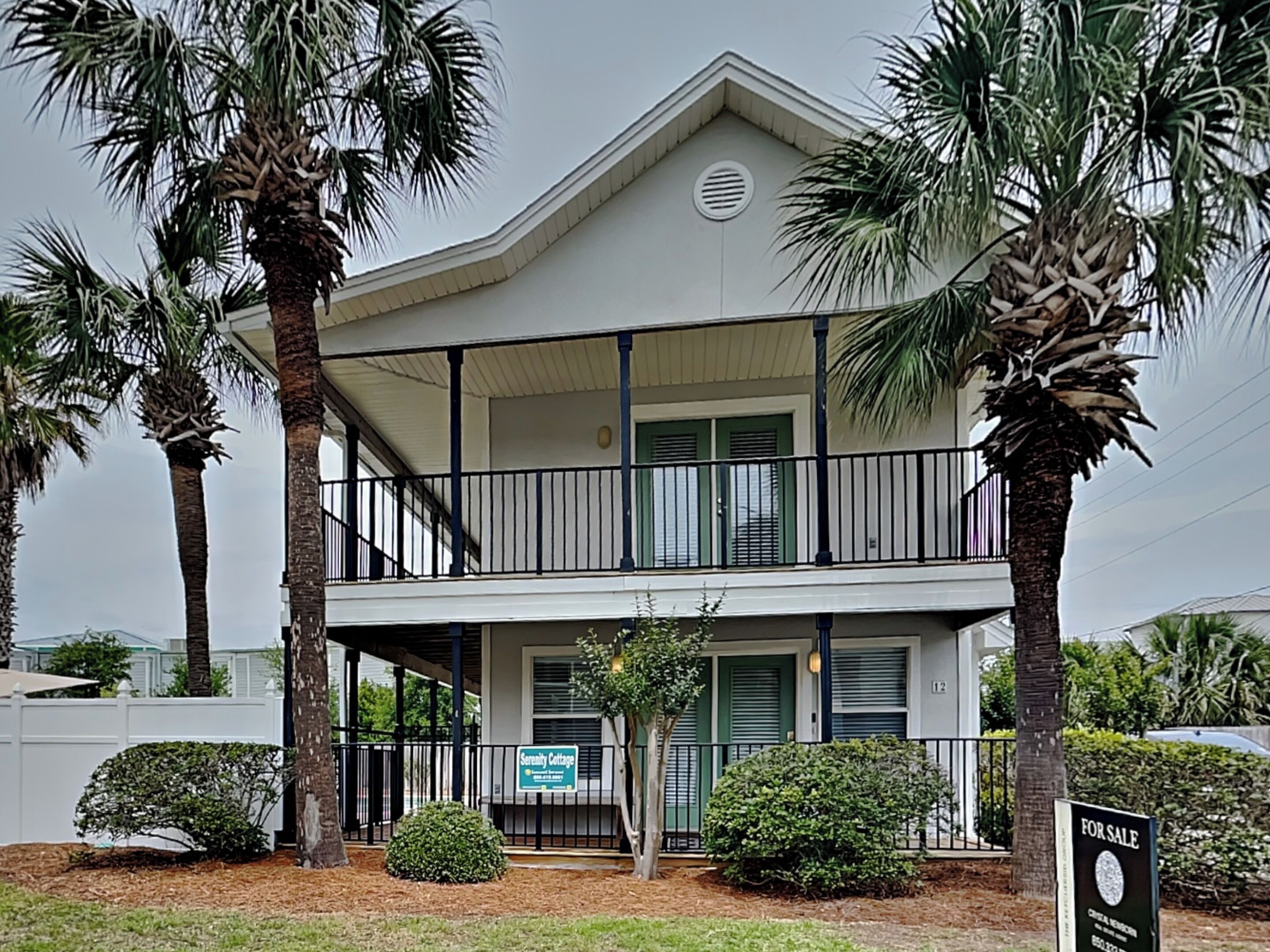 A two-story building with a balcony, surrounded by palm trees and a 'For Sale' sign.