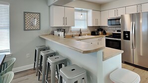 Een moderne keuken met witte kastjes, een roestvrijstalen koelkast, een magnetron en een spoelbak. Drie krukken en een vuilnisbak zijn aanwezig.