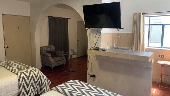 A compact hotel room with a bed, a small armchair, a TV mounted on the wall, a kitchenette with a sink, and a window with curtains.