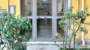 Une entrée d’immeuble avec une porte en verre, une poignée en métal et une petite fenêtre. Des plantes sont disposées devant la porte.