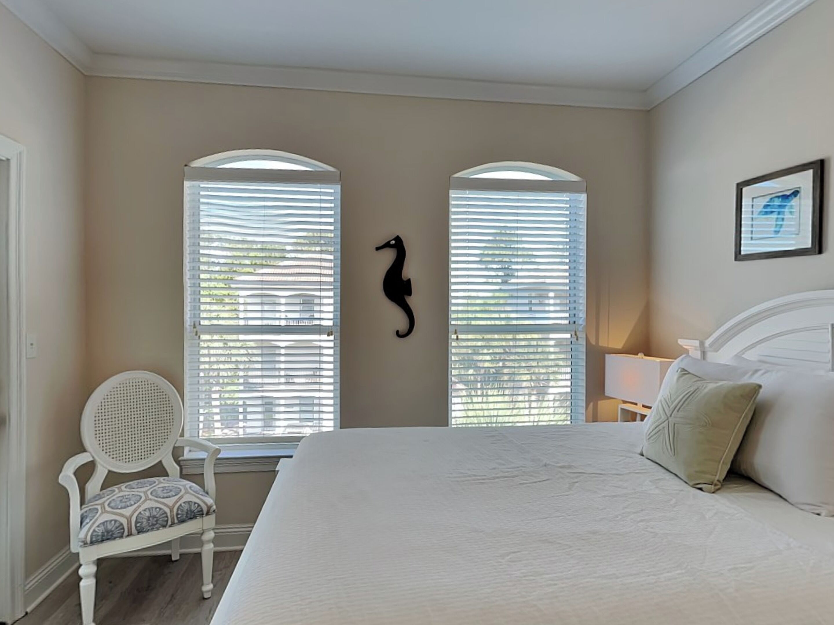 A bedroom with a large bed, two windows with blinds, a chair, a seahorse decoration, and a framed picture on the wall.