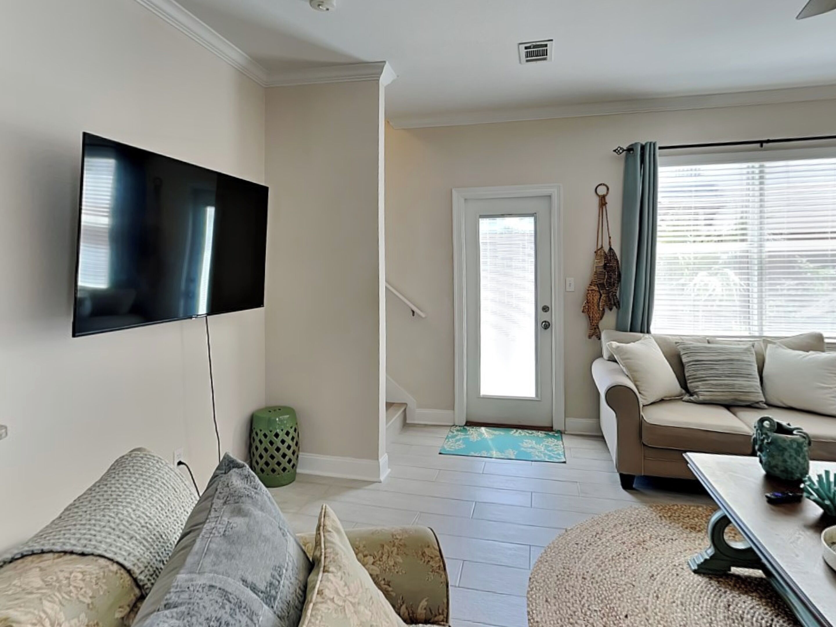 A living room with a flat-screen TV mounted on the wall, a beige sofa, a patterned rug, and a staircase leading to another level.