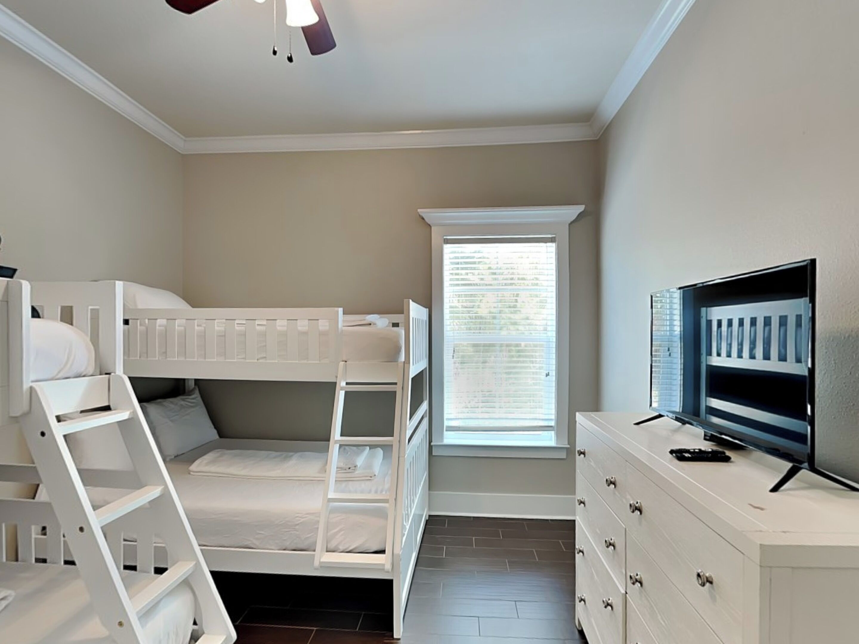 Une chambre avec un lit superposé, une commode, un téléviseur et une fenêtre avec des stores.