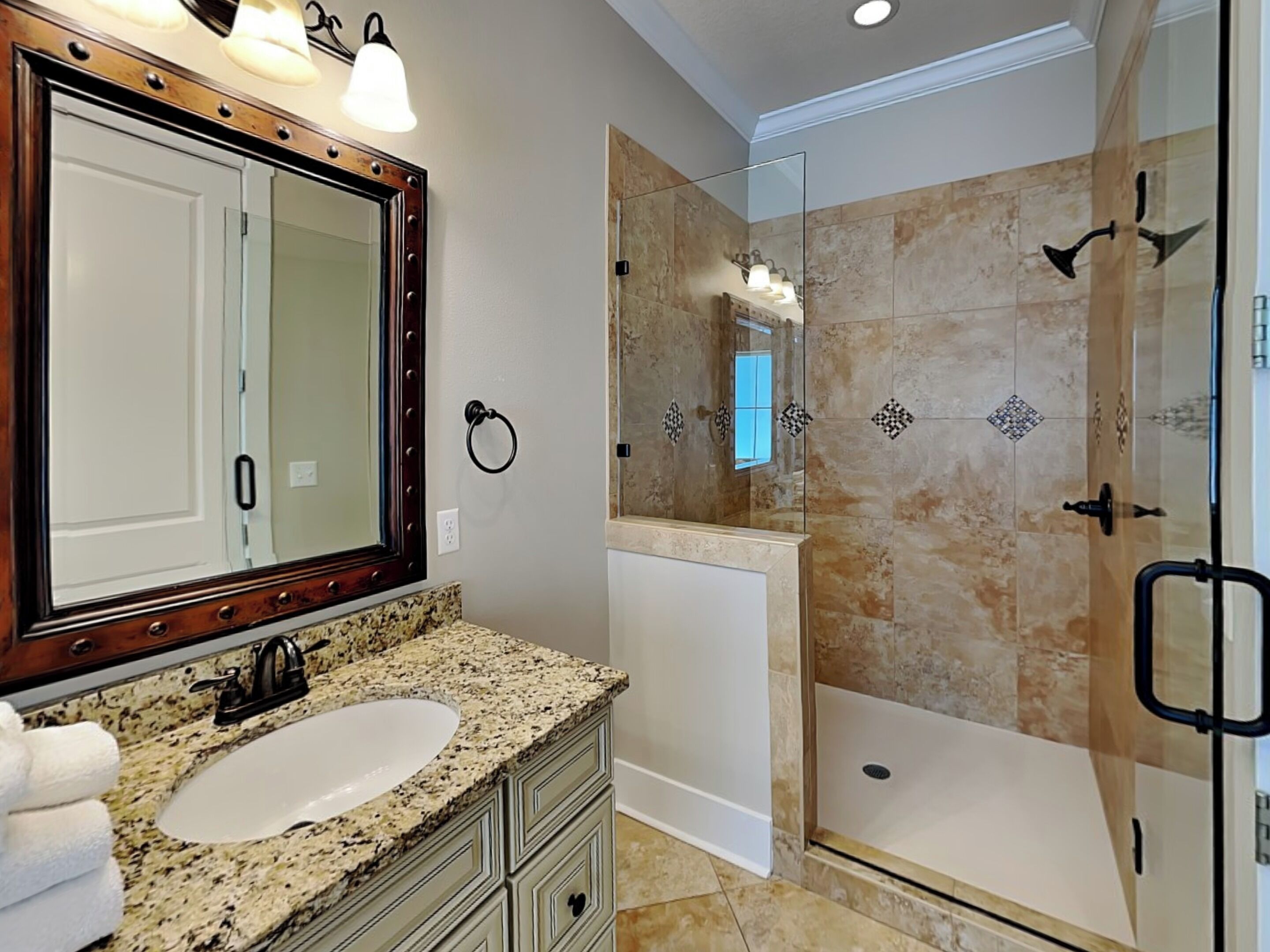 A bathroom with a granite countertop sink, a large mirror with lights, a towel rack, and a shower area with a glass door.