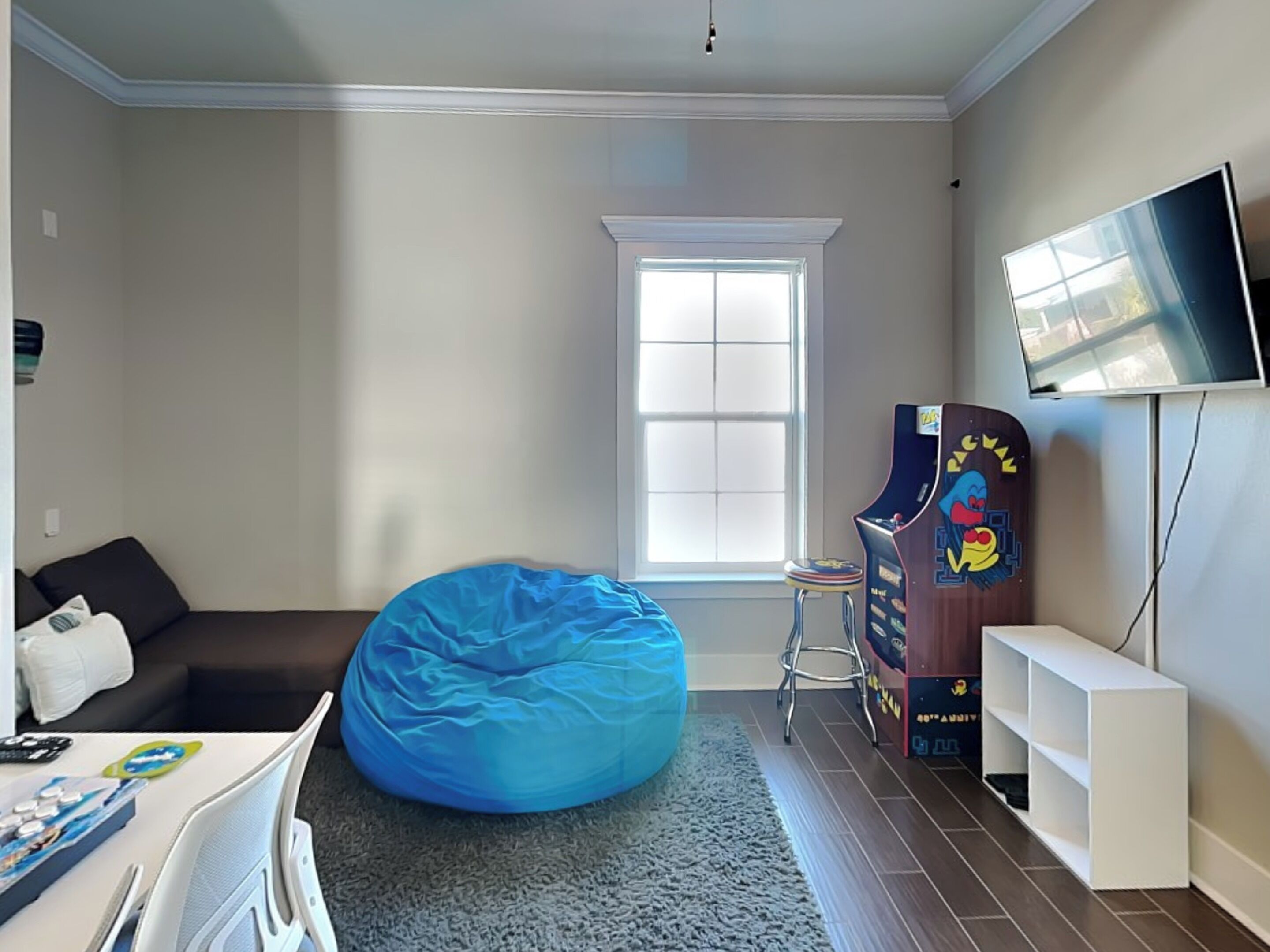 Una habitación con un sillón de peluche azul, un taburete pequeño, un televisor en la pared y una ventana.