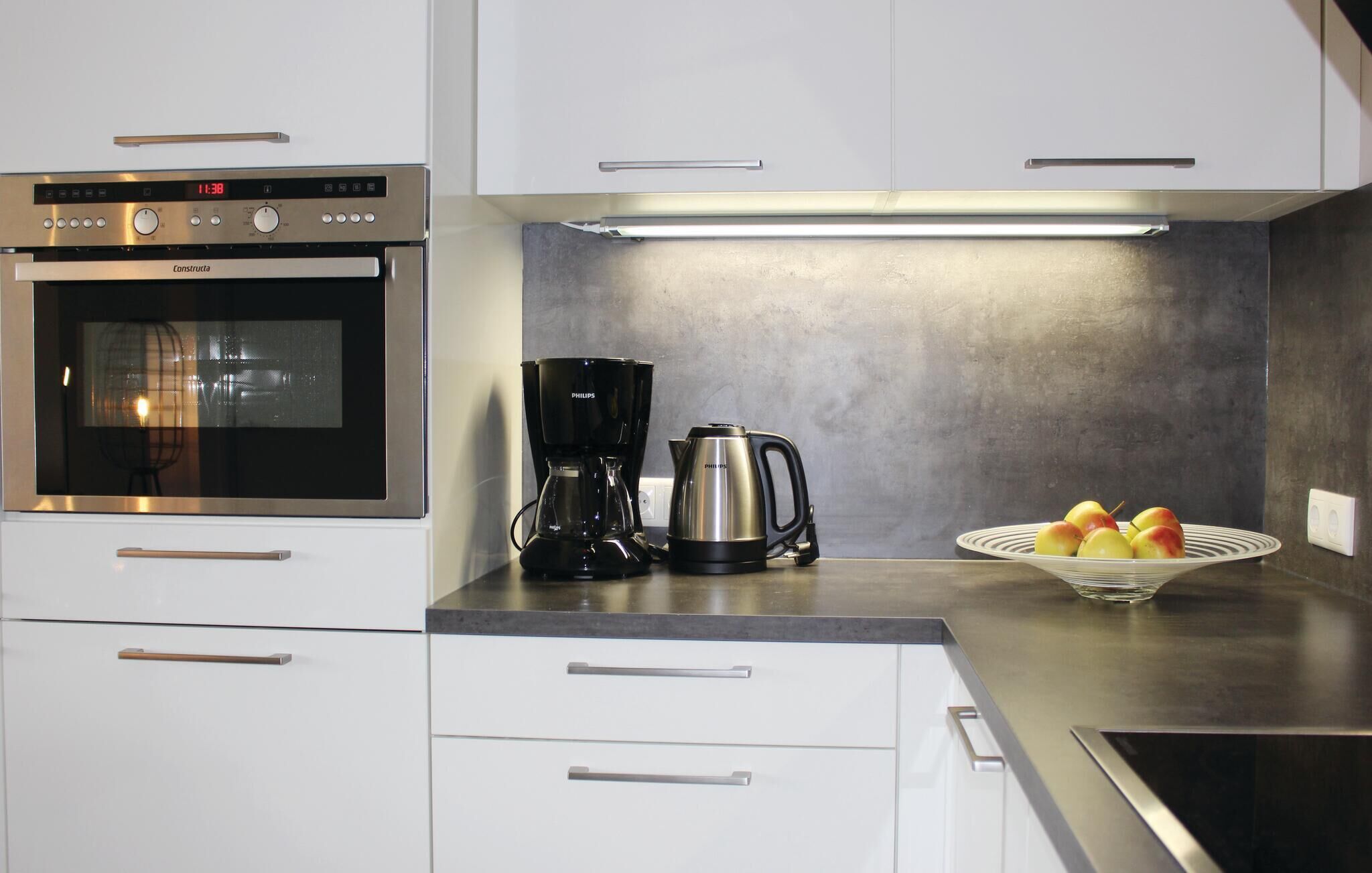 Una cocina moderna con horno integrado, cafetera y hervidor. Hay armarios y una encimera con un frutero.