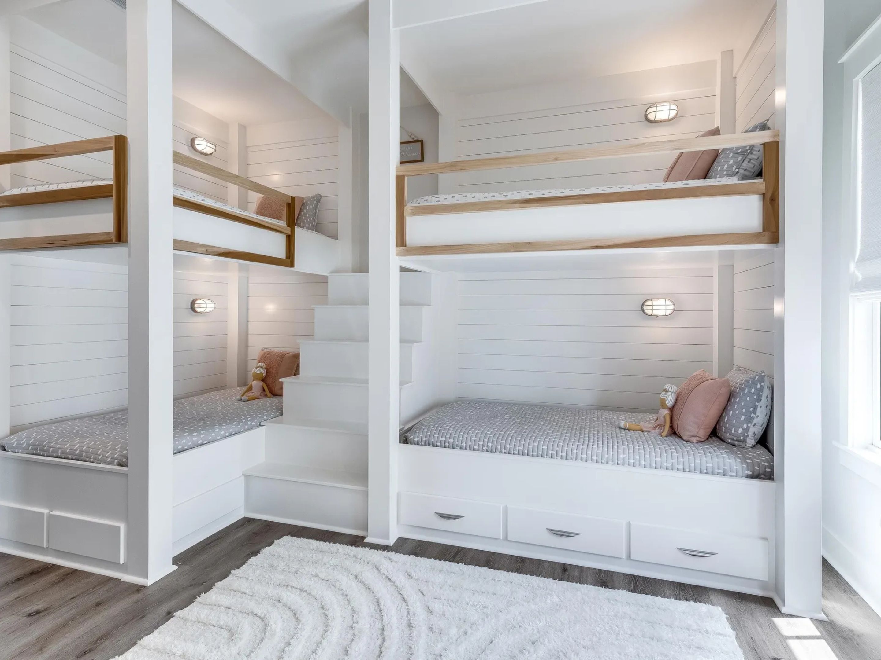 A children's bunk bed with a built-in desk and shelving, white walls, and a fluffy white rug.