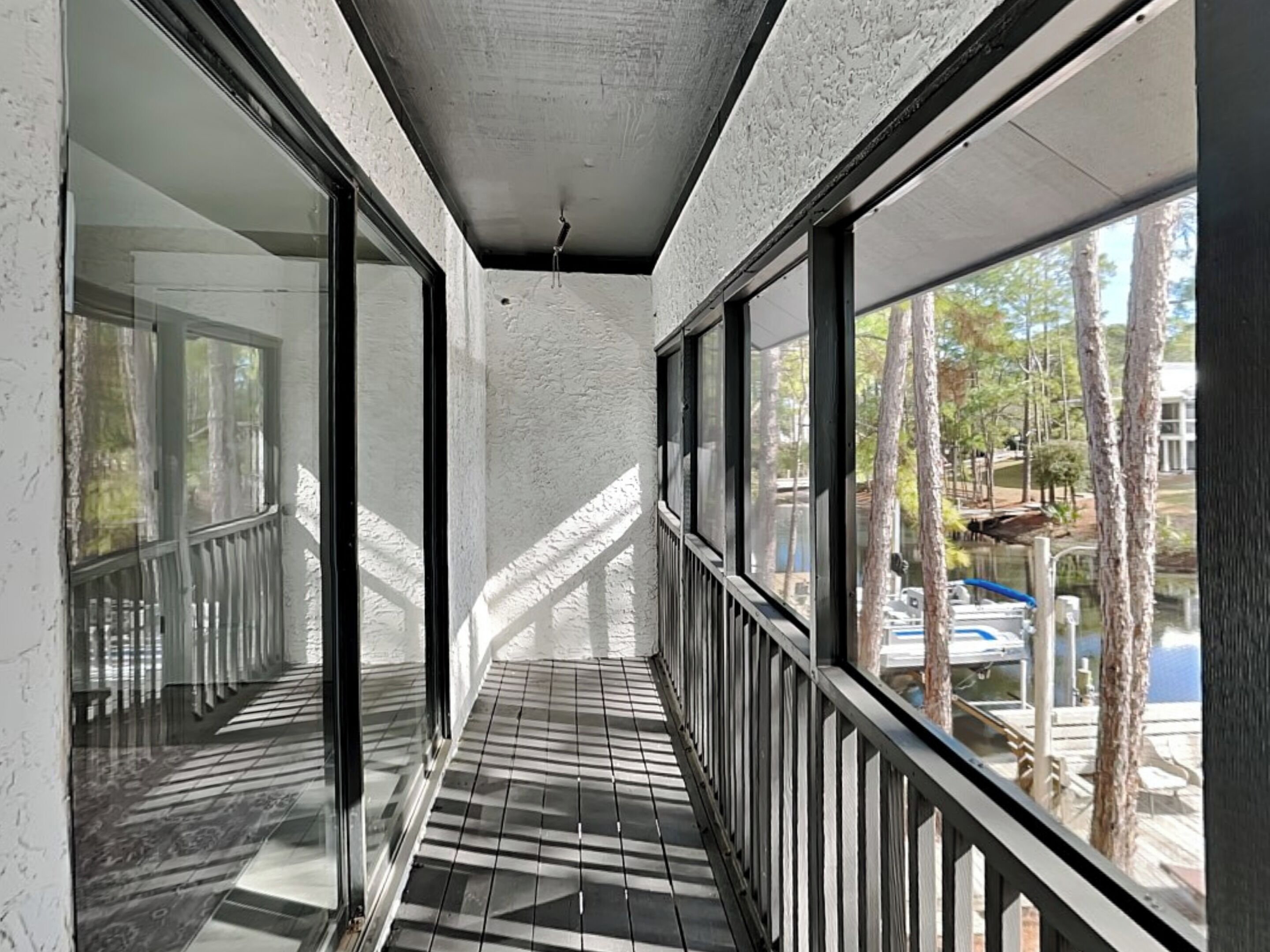 A corridor with large windows and a tiled floor, leading to a balcony with a view of trees and a dock.
