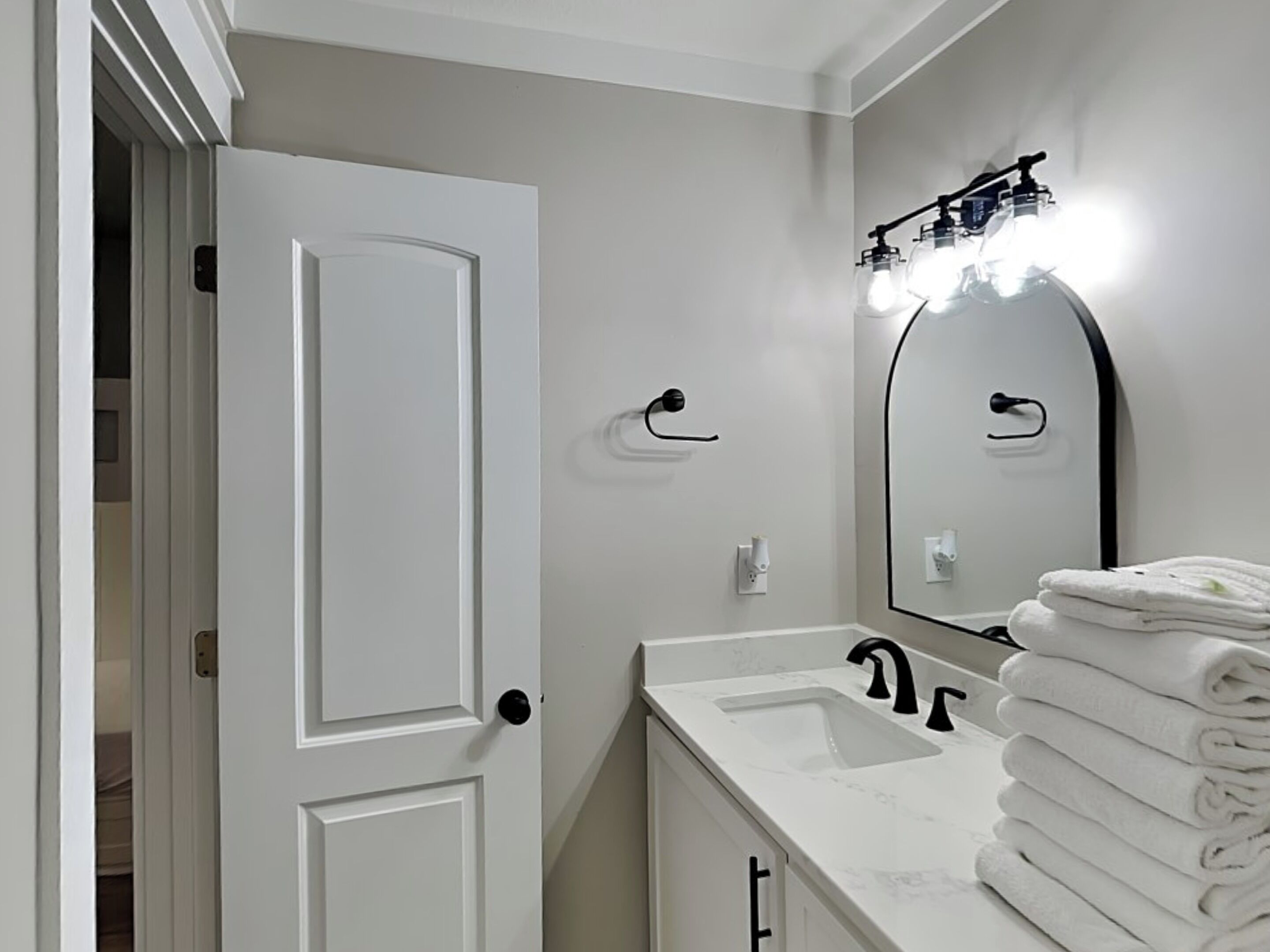 A bathroom with a white door, a sink with a mirror above it, and a towel rack with folded towels.