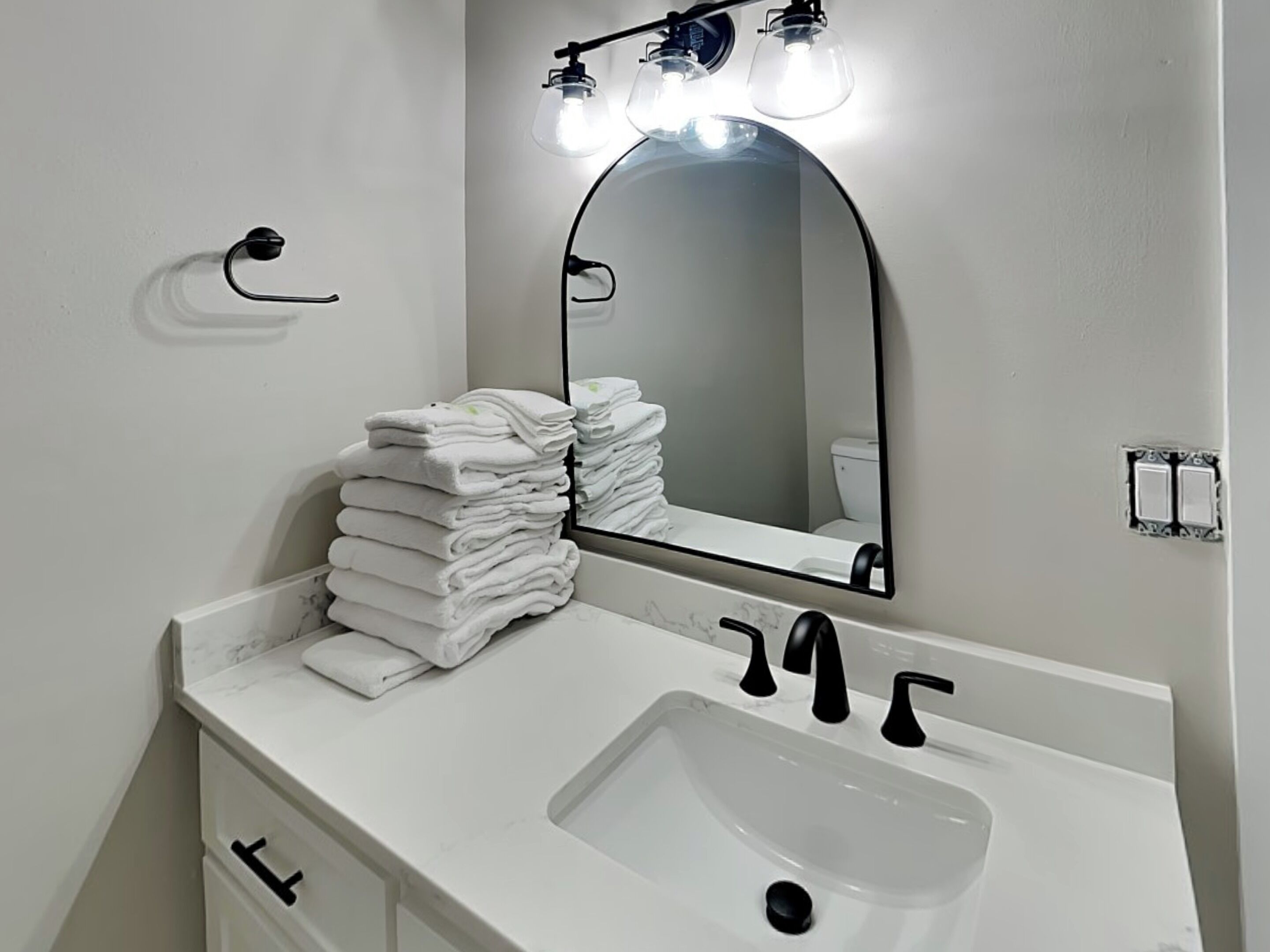 A bathroom with a white sink, a mirror with lights, and a stack of white towels.