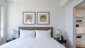 Ein modernes Hotelzimmer mit einem großen Bett, zwei Nachttischen mit Lampen, einem Kleiderschrank und zwei gerahmten Kunstwerken an der Wand.