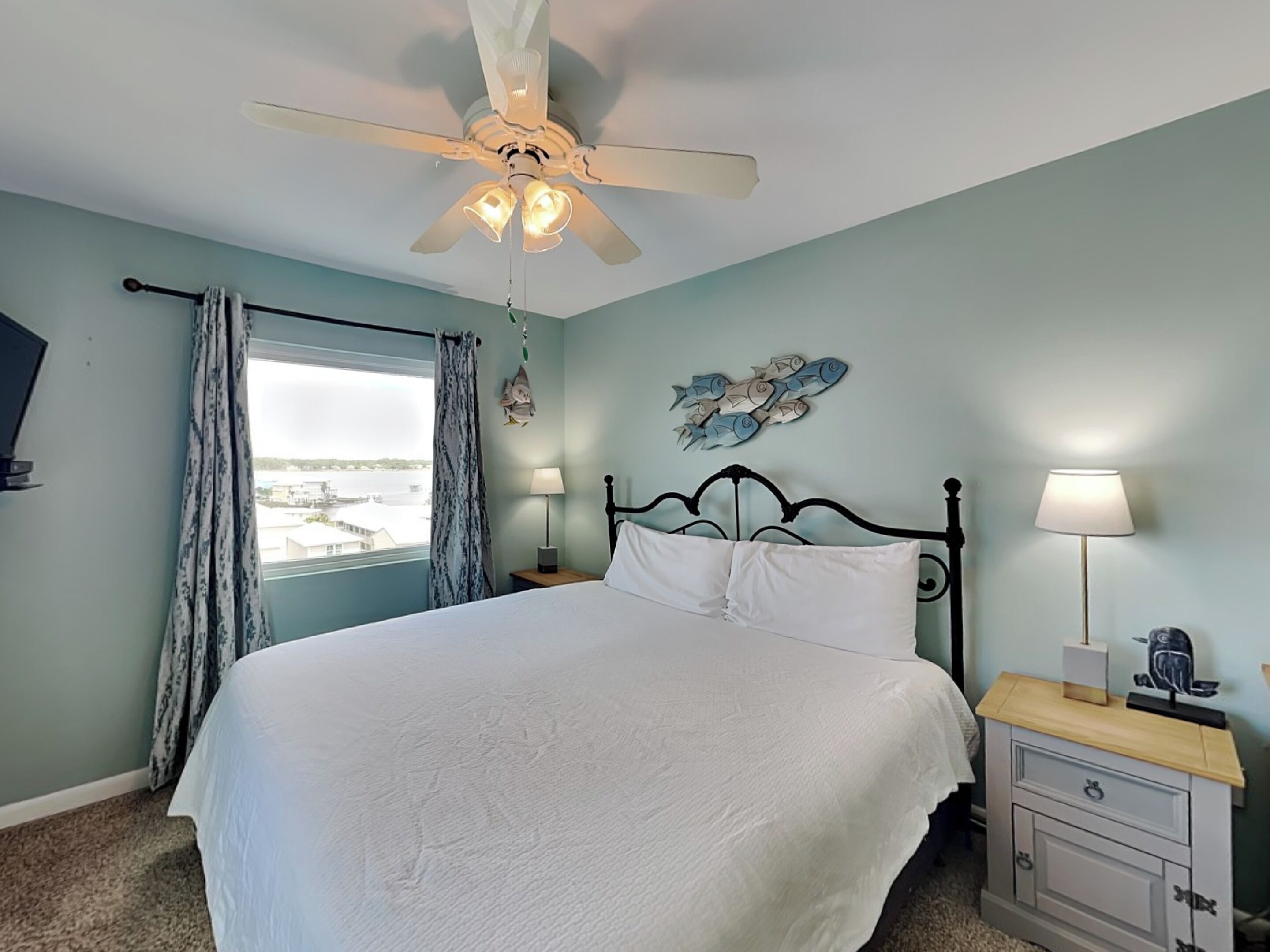 A bedroom with a bed, a ceiling fan, a window with curtains, a nightstand with a lamp, and decorative fish wall art.