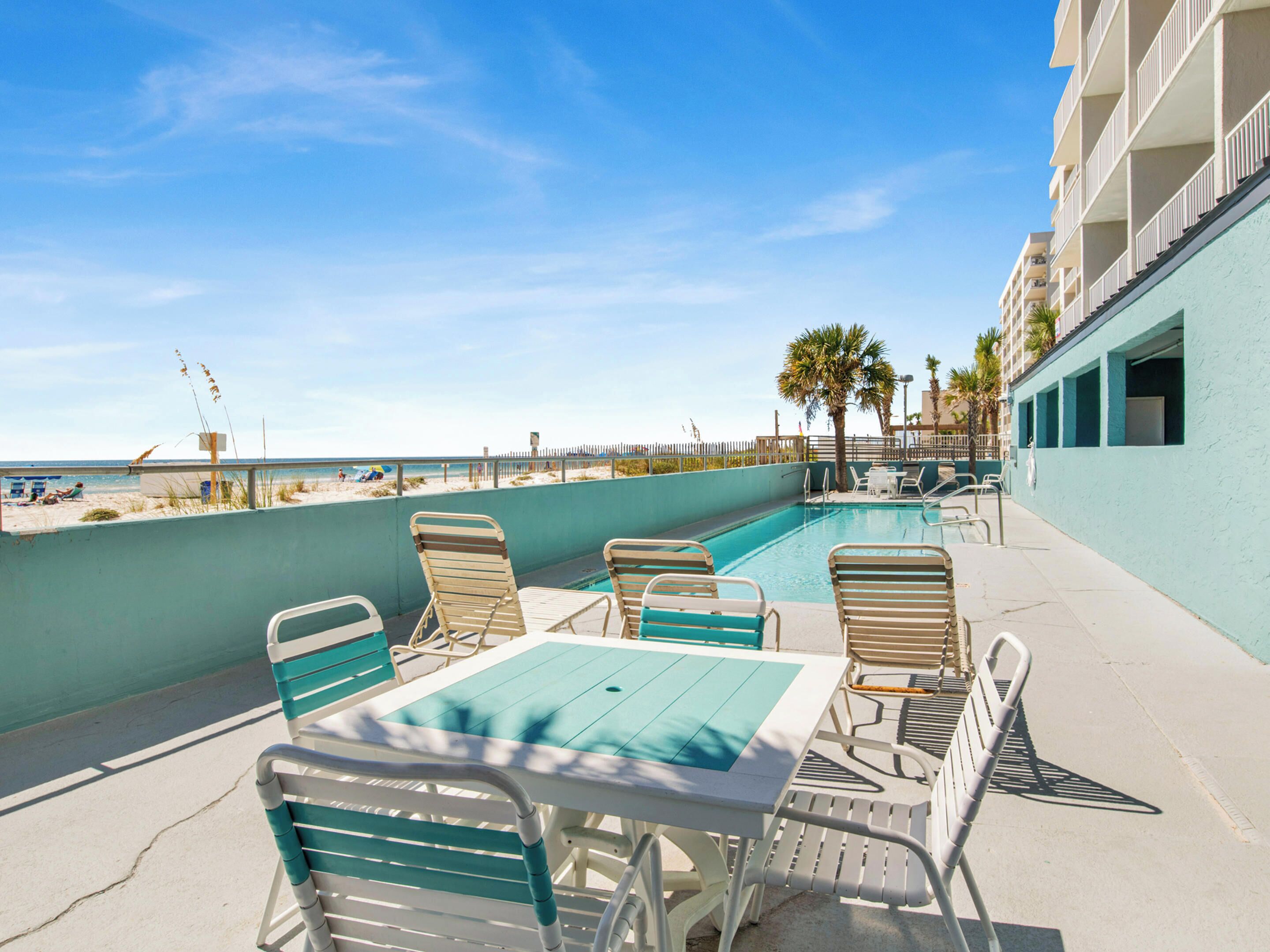 An outdoor pool area with lounge chairs and a table, overlooking a beach with palm trees and a clear sky.
