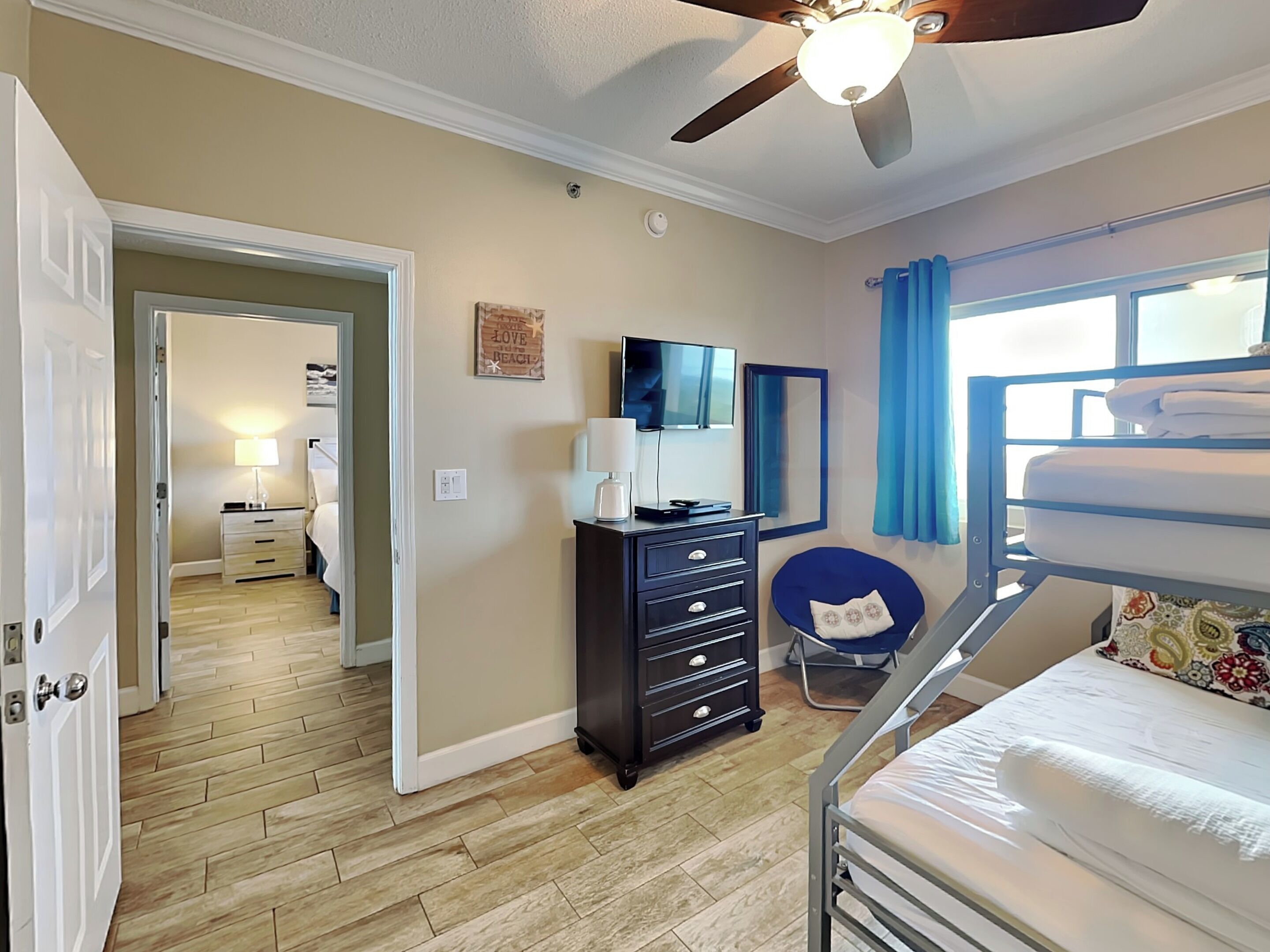 A bedroom with a bunk bed, a dresser, a desk with a chair, a ceiling fan, and a window with curtains.