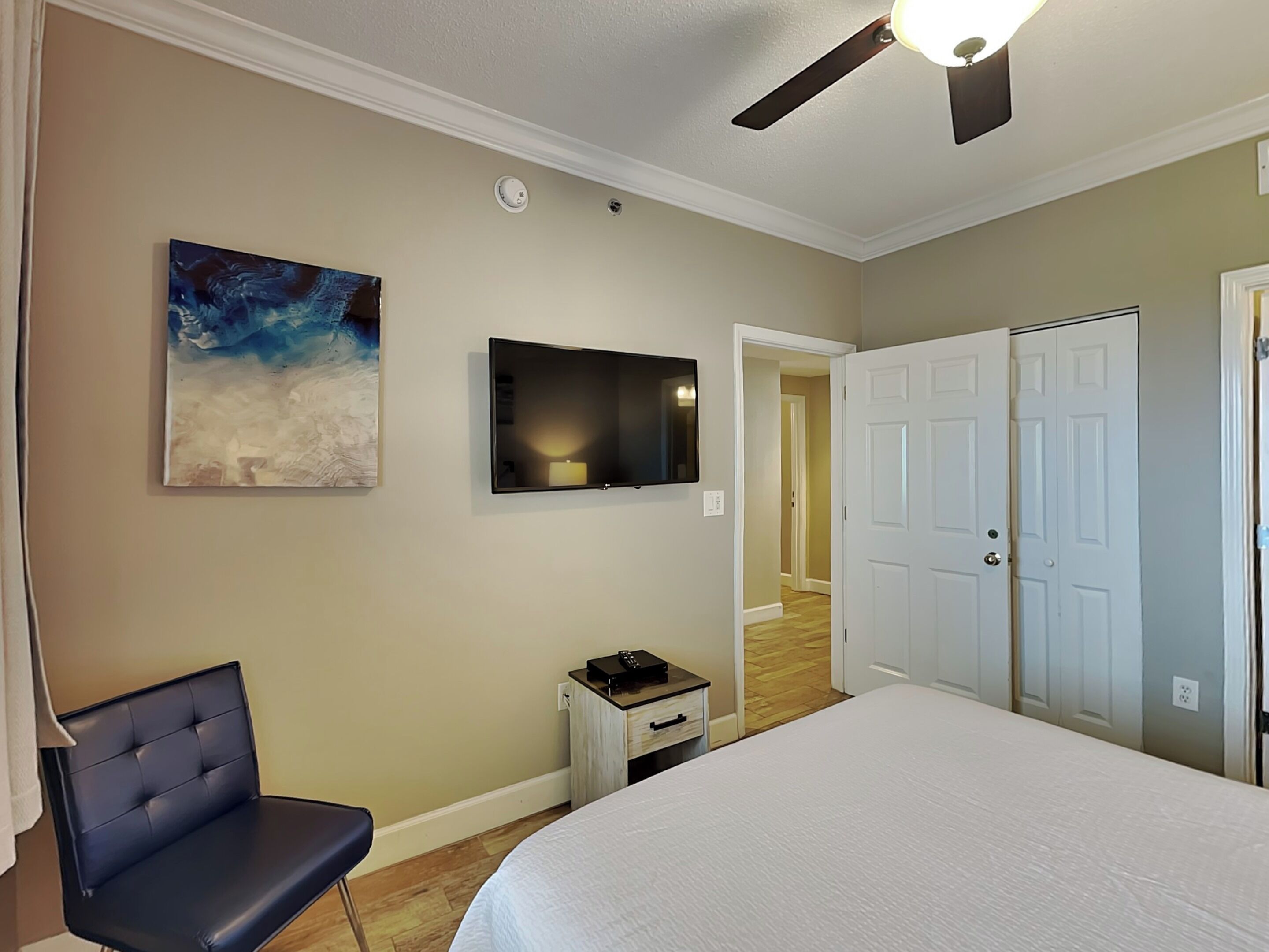A bedroom with a bed, a chair, a TV mounted on the wall, a ceiling fan, and a door leading to another room.
