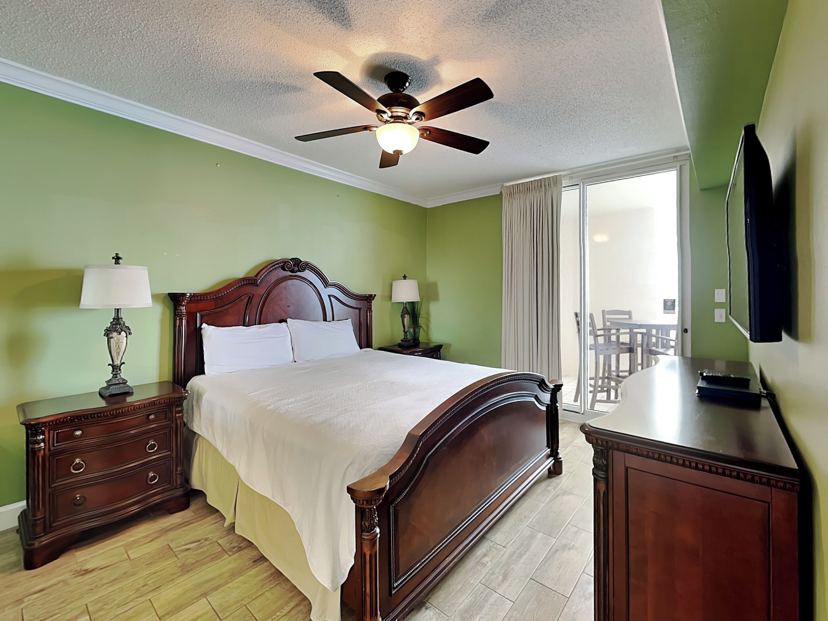 A hotel room with a wooden bed, two bedside tables with lamps, a ceiling fan, and a view of the outside balcony.