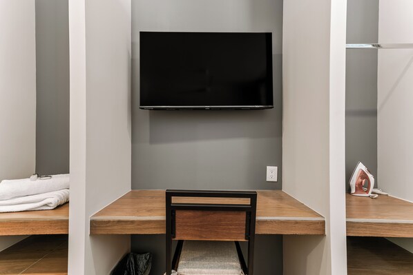A hotel room with a flat-screen TV mounted on the wall, a wooden desk with a chair, and a small ironing board with an iron.