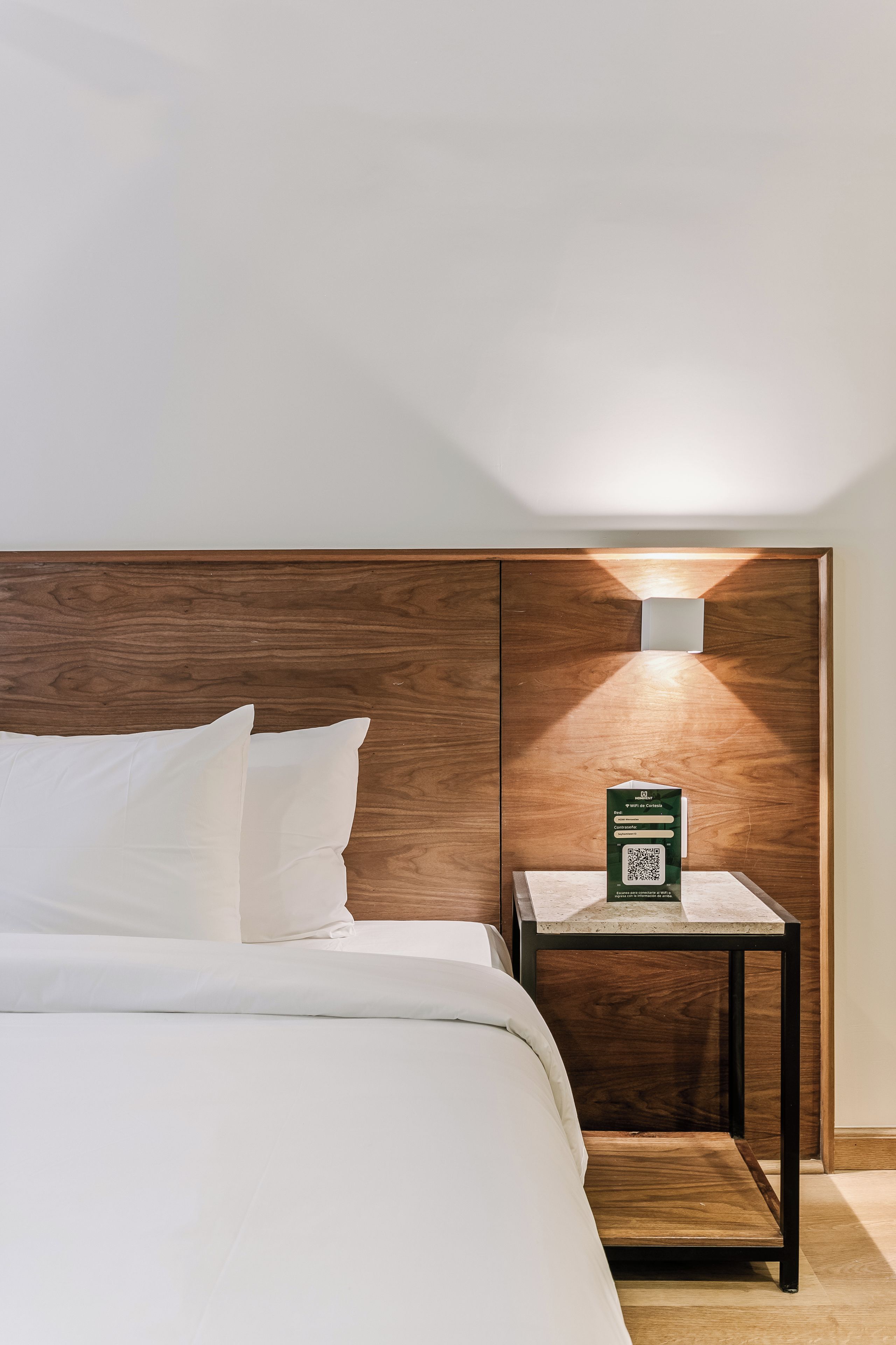A neatly made bed with white linens, a wooden headboard, and a small bedside table with a green sign and a QR code.