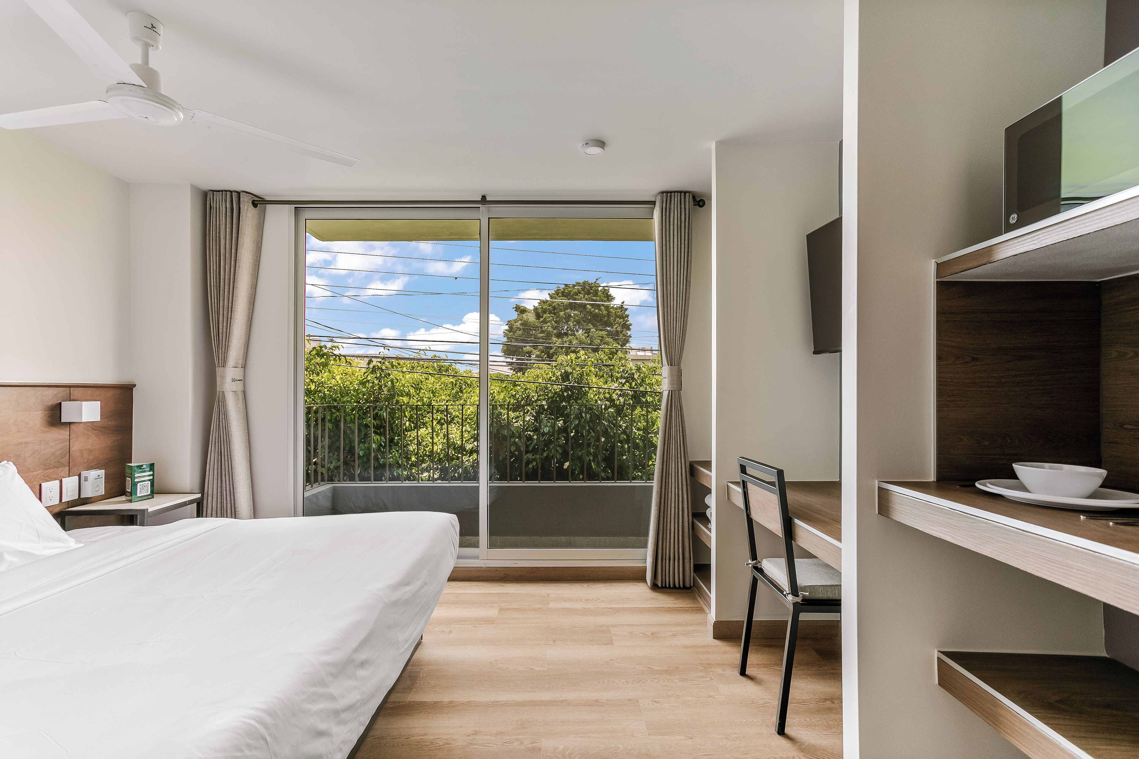 A hotel room with a bed, a desk, a chair, a TV, and a balcony with a view of greenery and a tree.