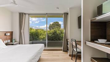 A hotel room with a bed, a desk, a chair, a TV, and a balcony with a view of greenery and a tree.