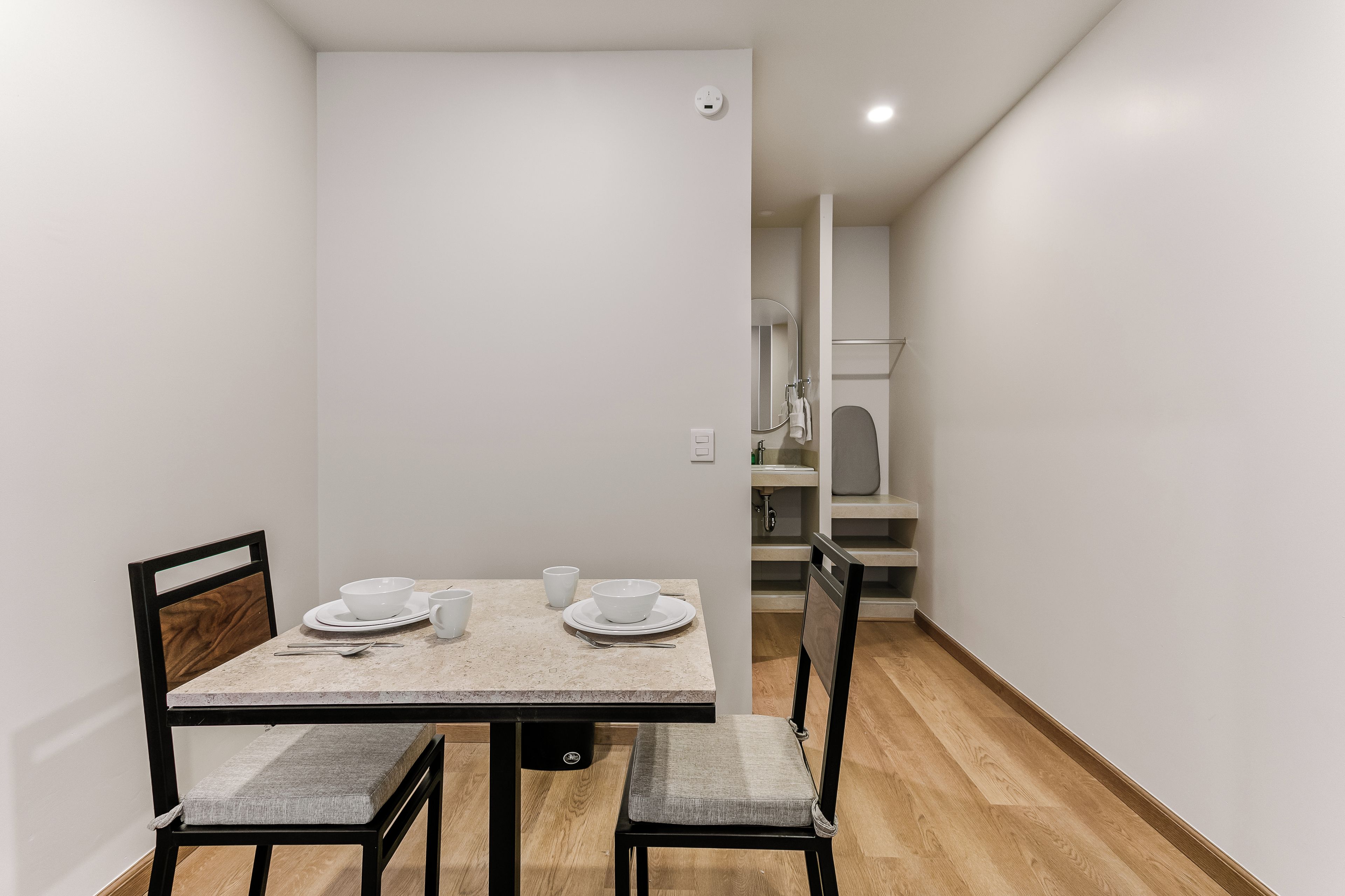 A dining area with a table set for two, chairs, a mirror, and a view into the kitchen.