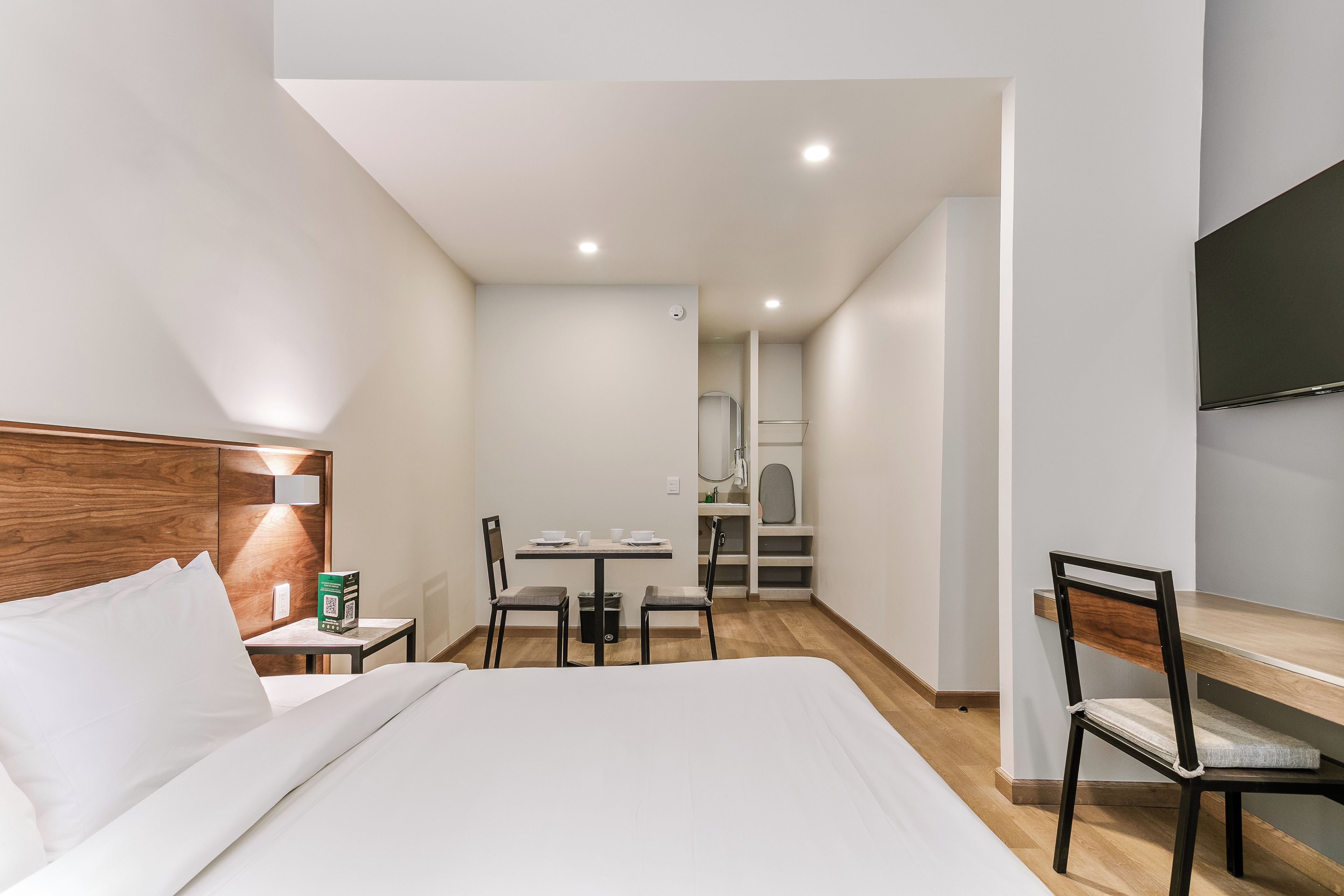 A hotel room with a bed, a desk, a chair, a small table with a chair, and a kitchenette in the background.