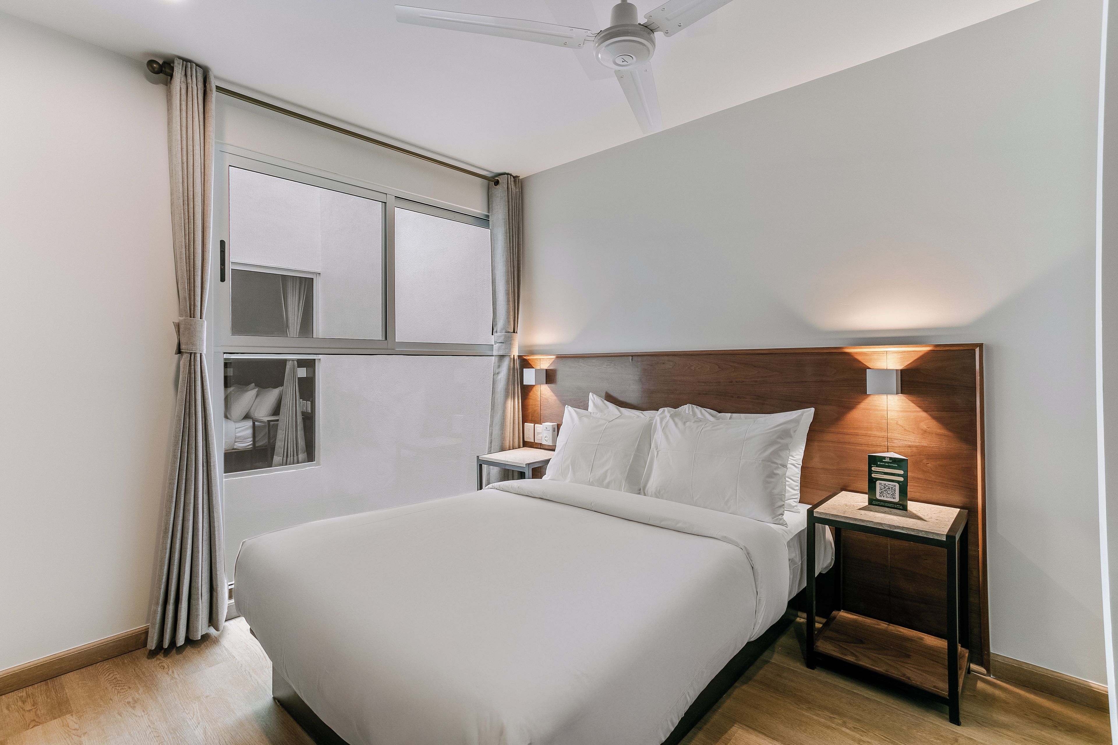 Una habitación de hotel con una cama grande, una mesita de noche con lámpara, una ventana con cortinas y un ventilador de techo.