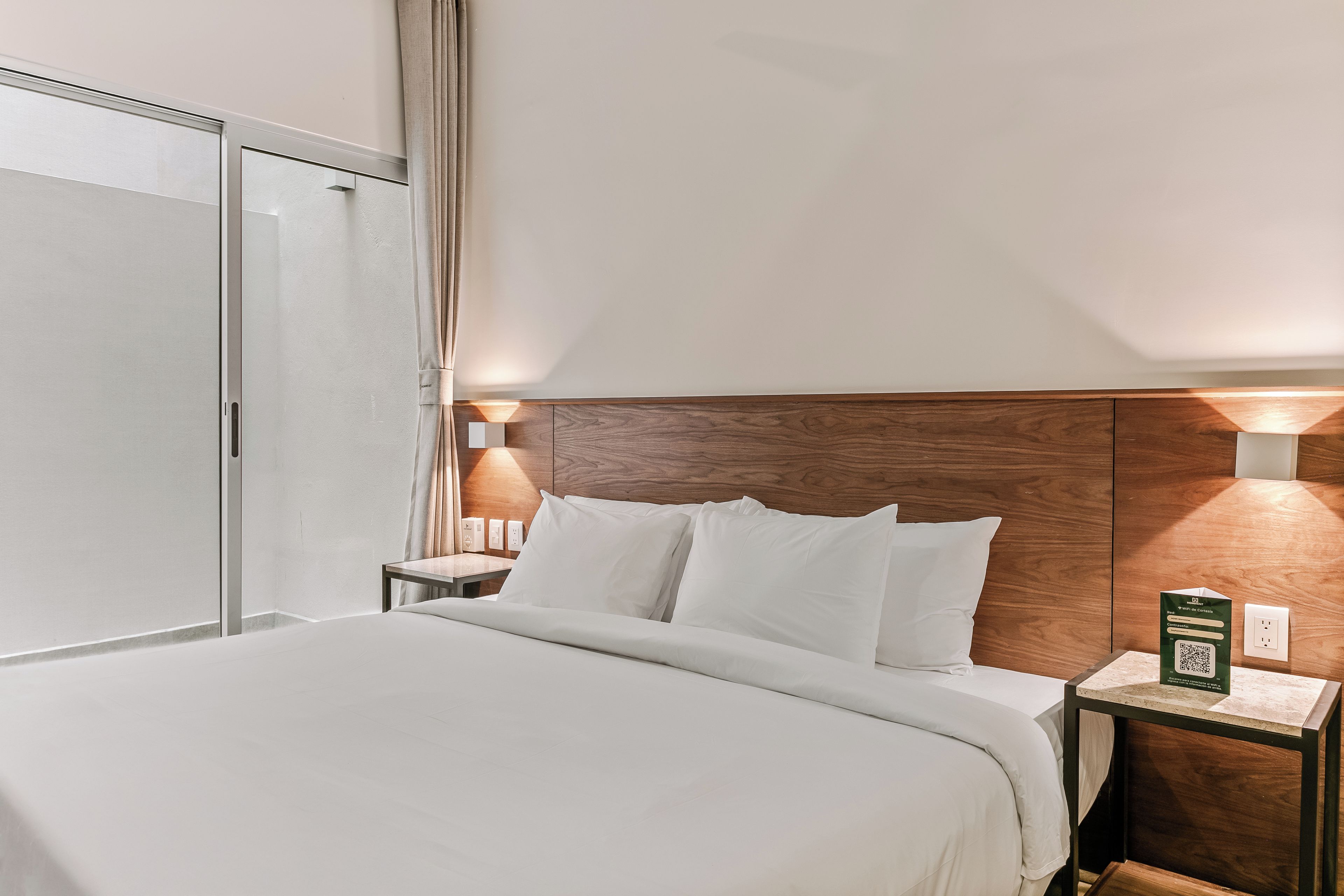 A hotel room with a bed, a nightstand, a wooden headboard, and a wall-mounted light fixture.