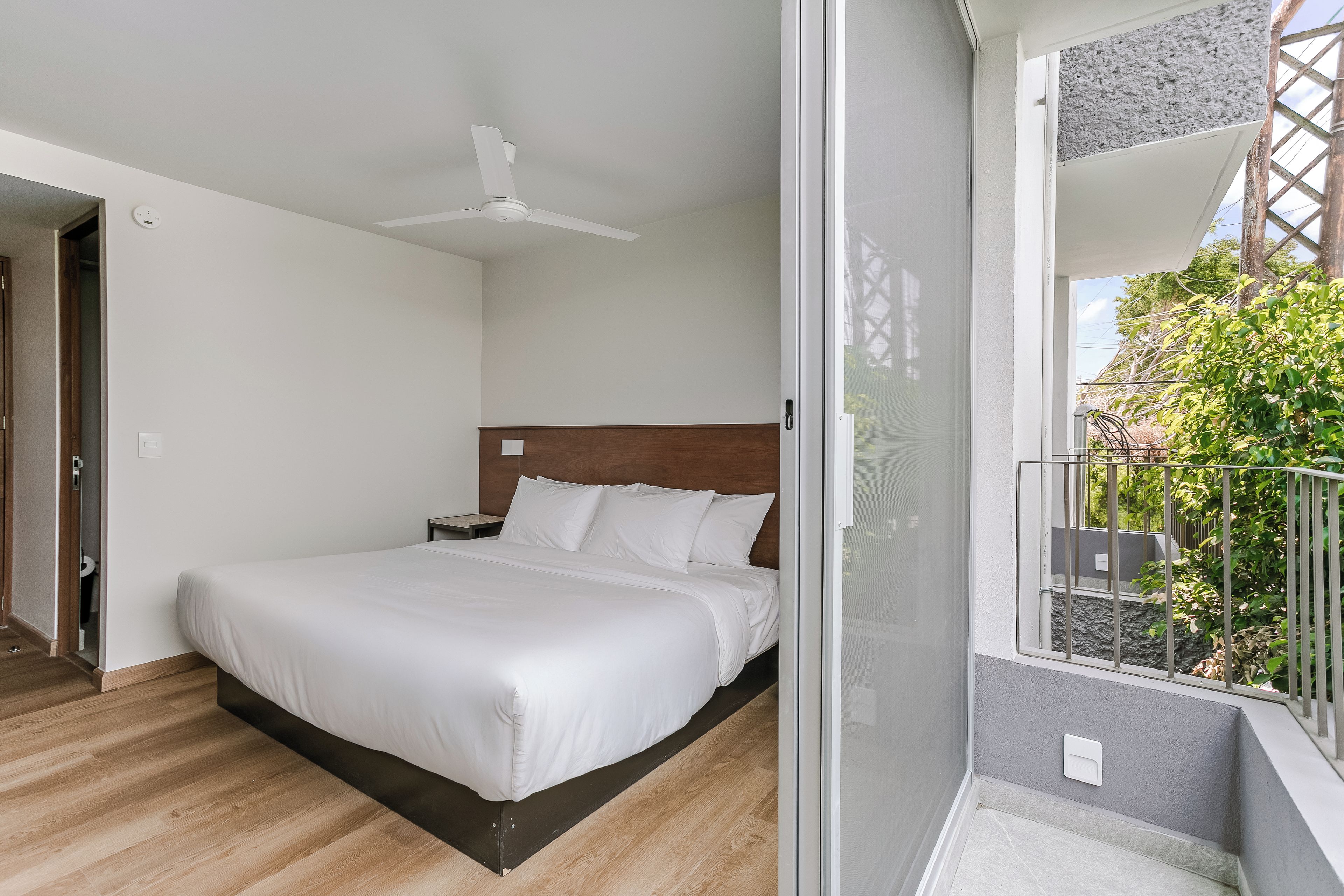 Habitación de hotel con cama, ventilador de techo, ventana con vista a zonas verdes y balcón con barandilla.