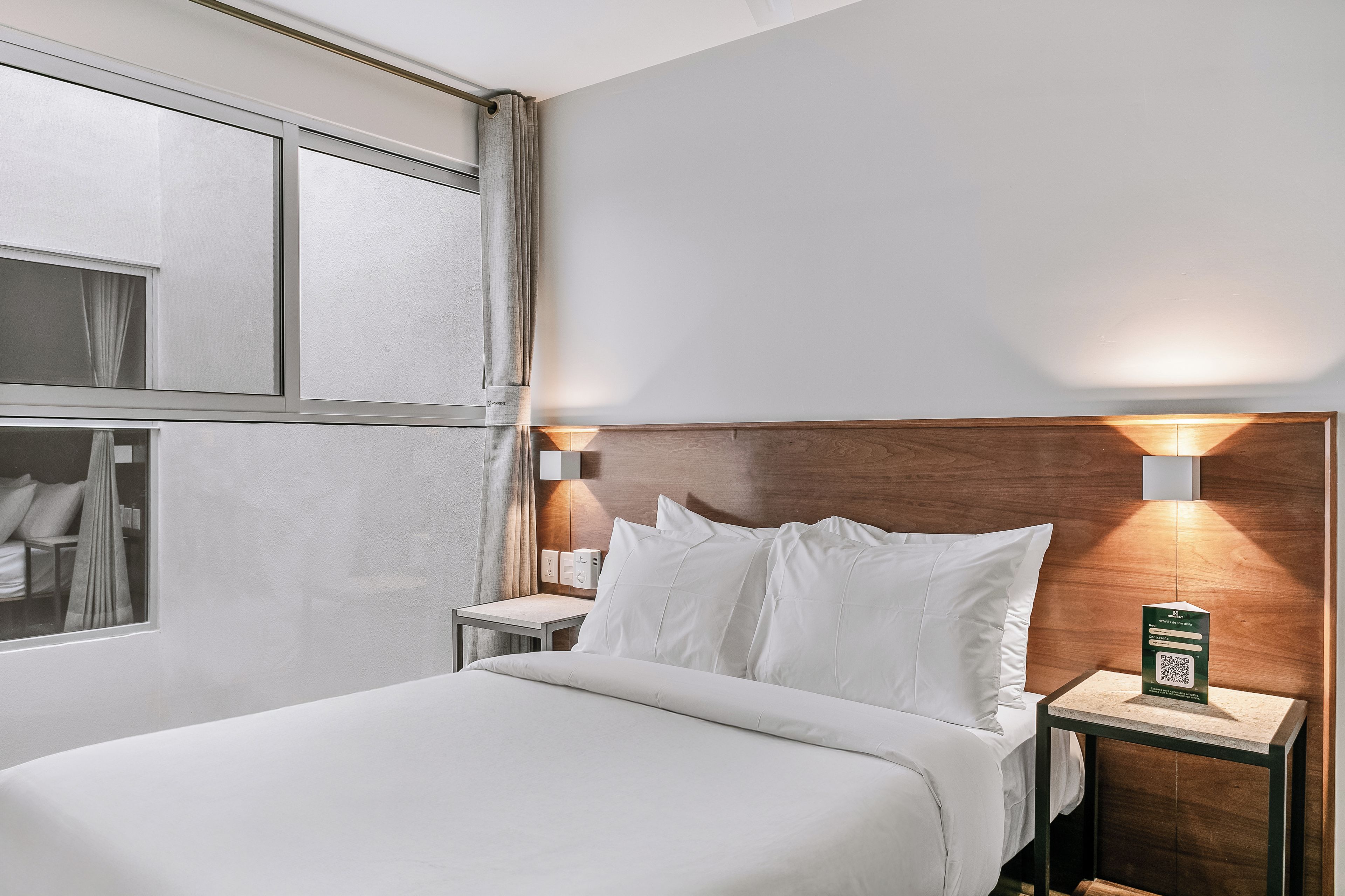 A hotel room with a bed, bedside tables, a nightstand, a wall-mounted light, and a QR code on the nightstand.