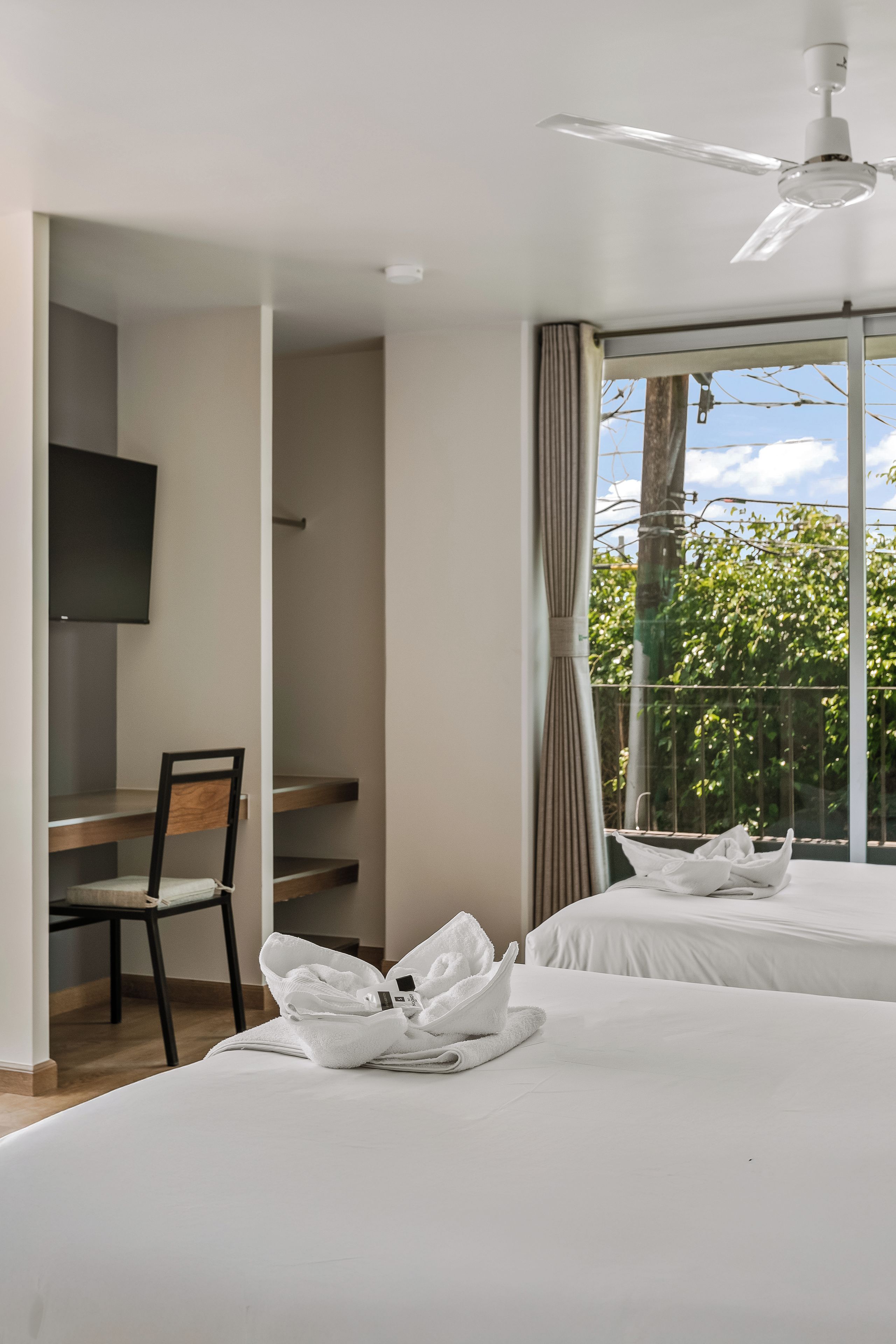 Une chambre d’hôtel avec deux lits, un bureau, une chaise, une télévision et un balcon donnant sur la verdure.