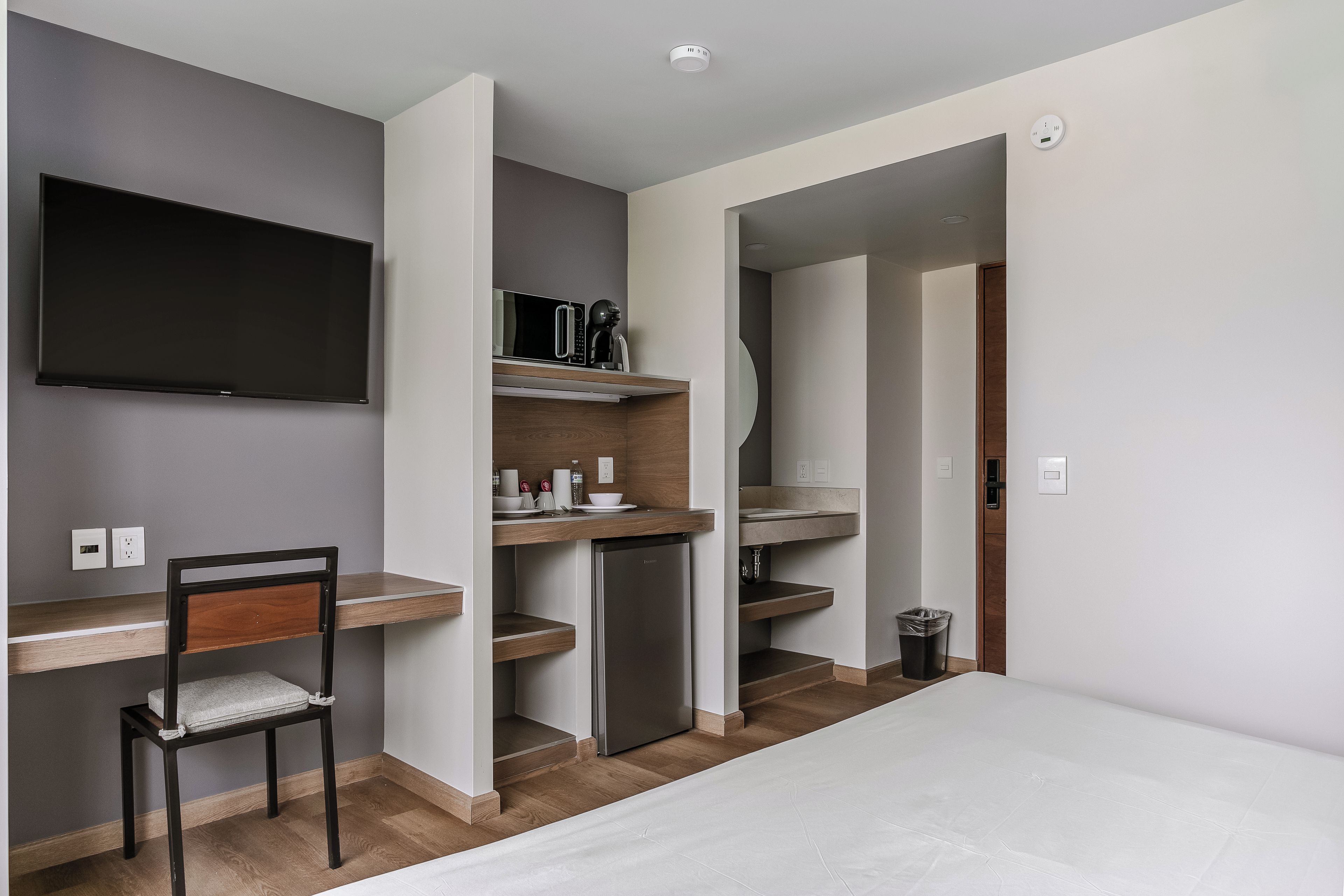 Une chambre d’hôtel moderne équipée d’un téléviseur à écran plat, d’un bureau en bois, d’une chaise, d’un mini-réfrigérateur, d’un lavabo intégré et d’une poubelle.