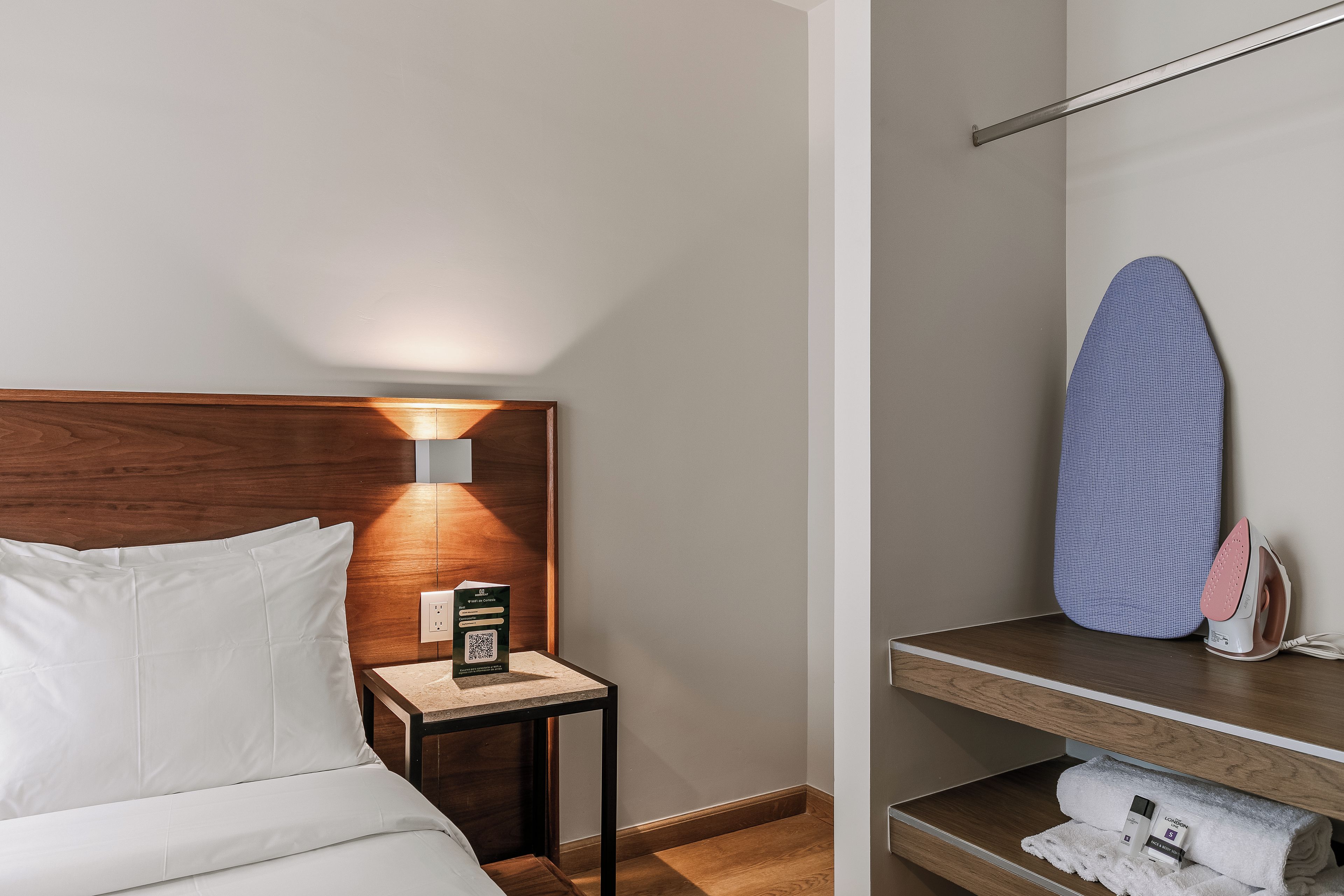 A hotel room with a bed, a nightstand with a lamp, a mirror, and a shelf with a towel and an iron.