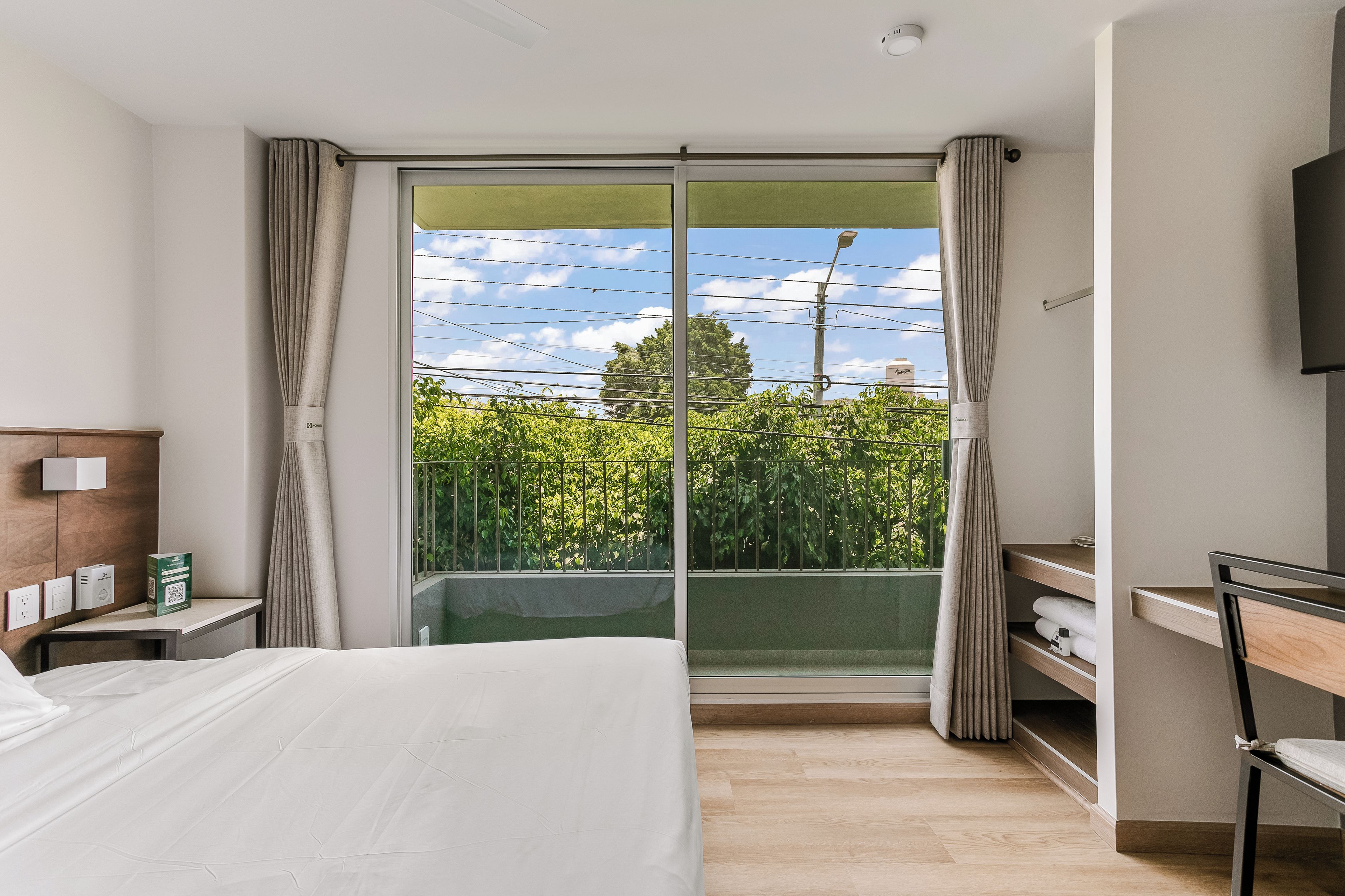 A hotel room with a bed, a desk, a chair, a TV, and a sliding glass door to a balcony with a view of greenery and a clear sky.