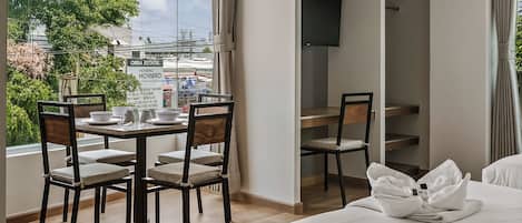 A hotel room with a bed, a dining table with chairs, a TV mounted on the wall, and a view of the city through the window.