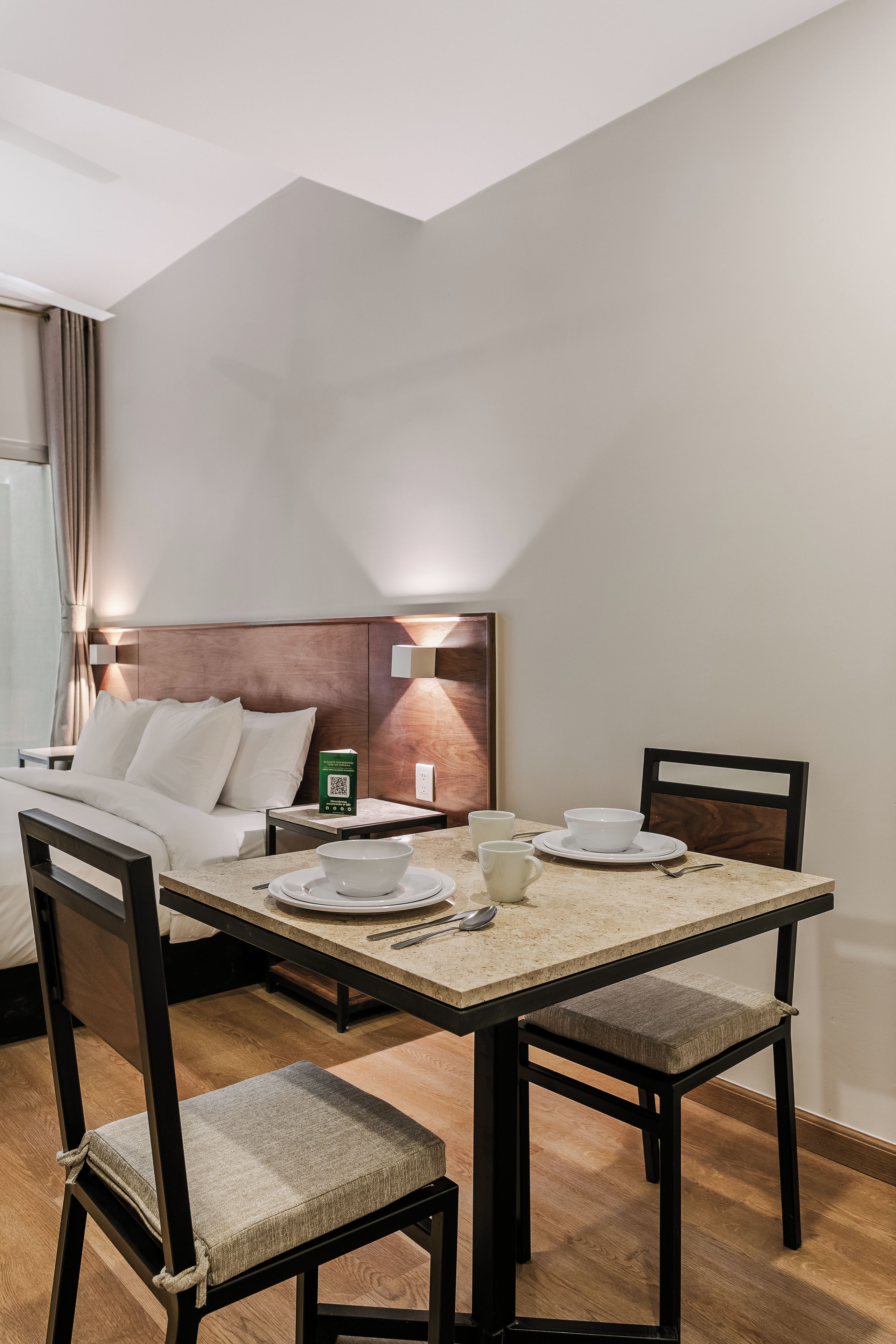 A hotel room with a dining table set for two, a bed with white linens, and a headboard with a light fixture.