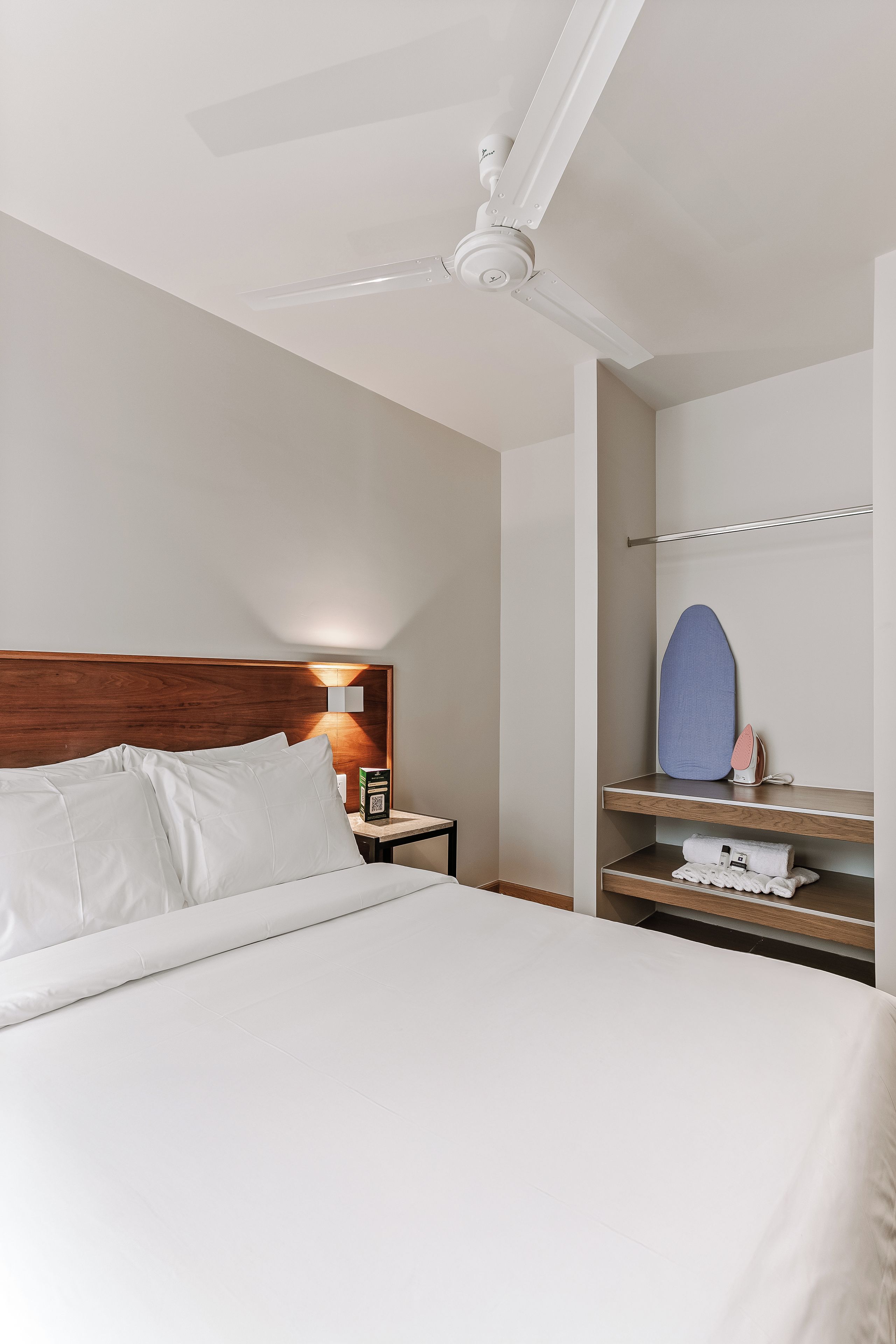 A hotel room with a bed, a nightstand, a mirror, and a shelf with a towel and an iron.