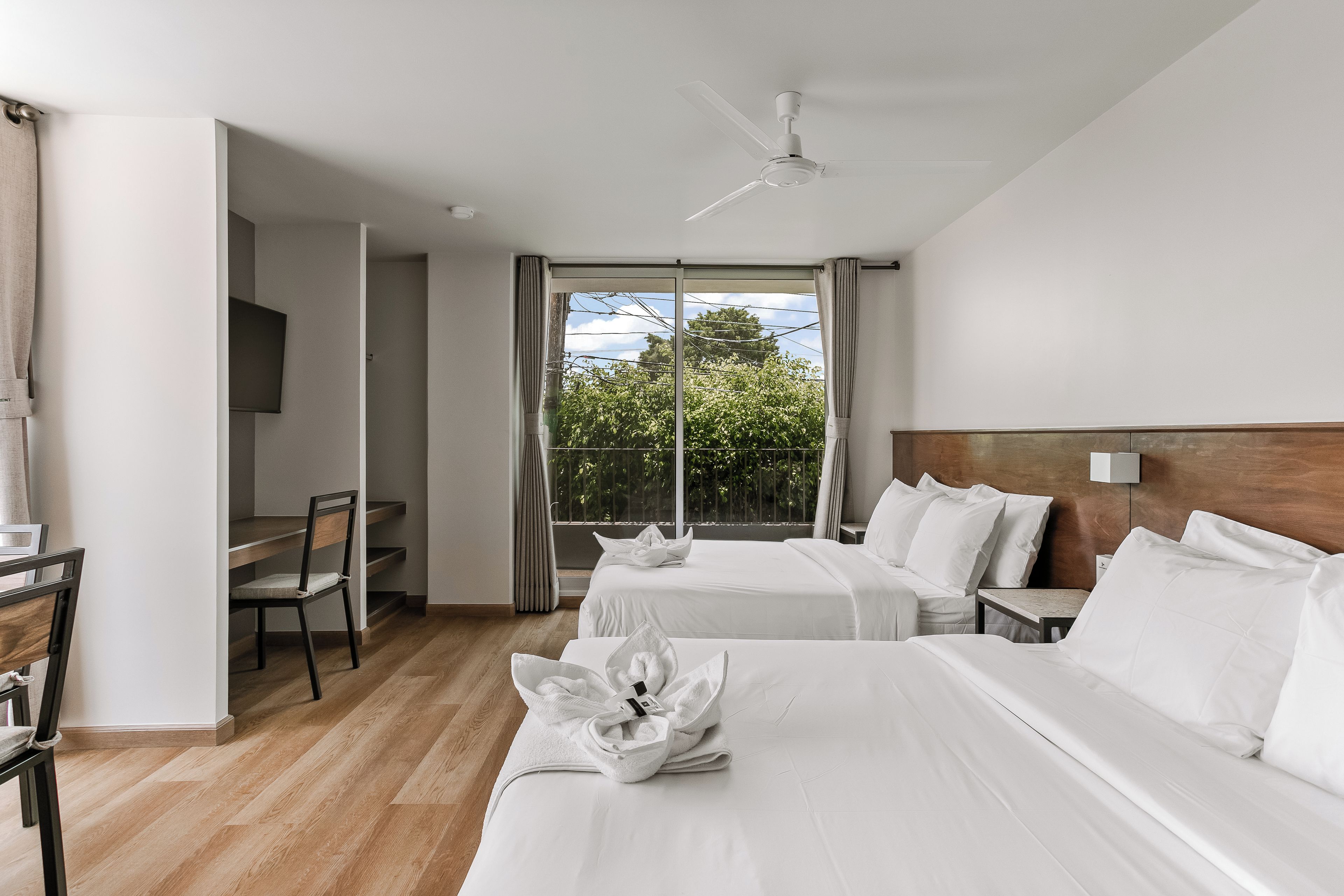 A hotel room with two beds, a desk, a chair, a TV, and a balcony with a view of trees.