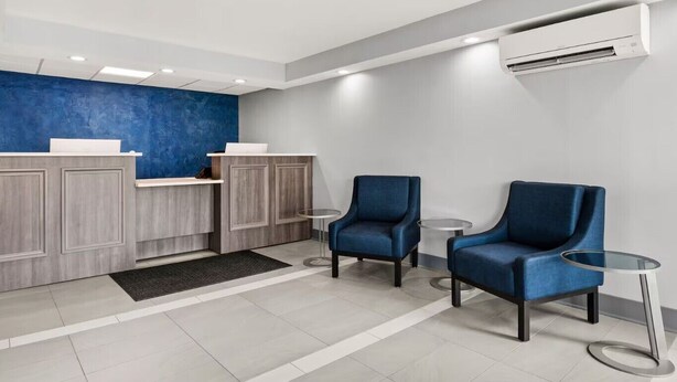 A reception area with a blue accent wall, two blue armchairs, a reception desk, and a small round table. - Baymont By Wyndham Batavia - Darien Lake (Elba)